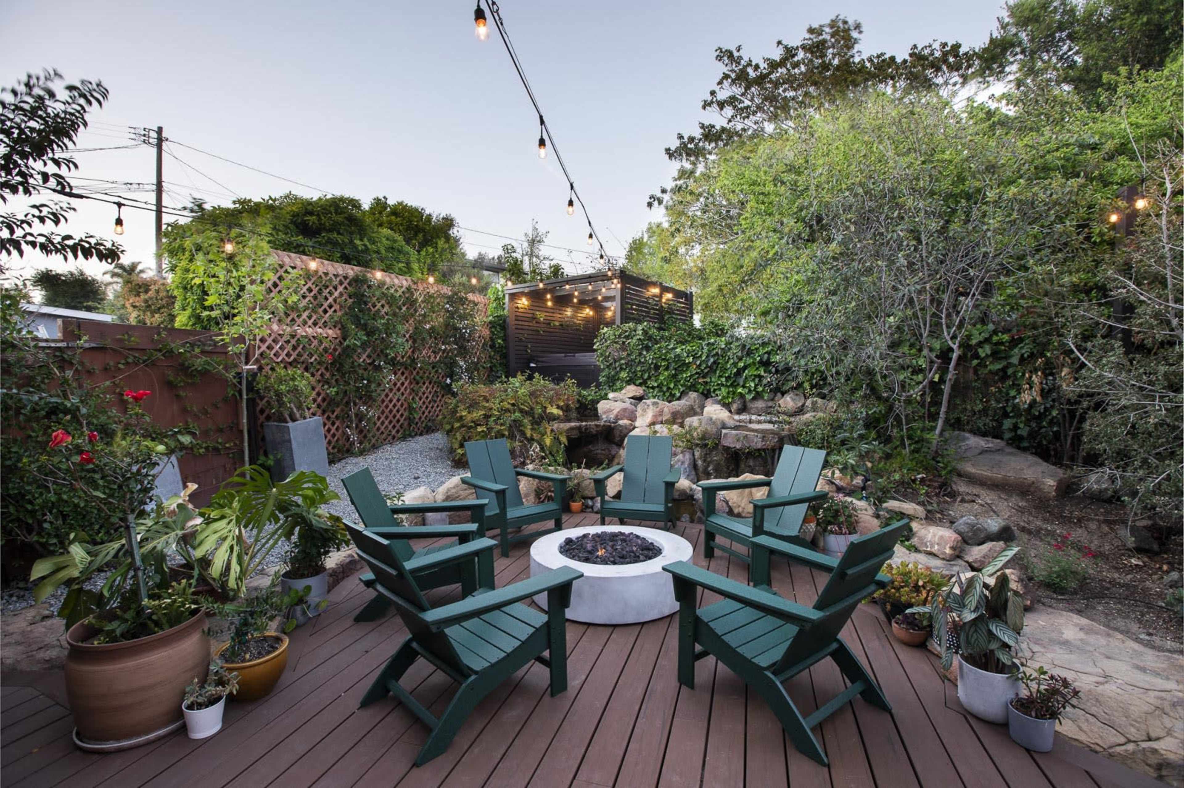 A circular seating area with six green Adirondack chairs surrounds a central fire pit, set within a well-landscaped backyard featuring plants and decorative stones.