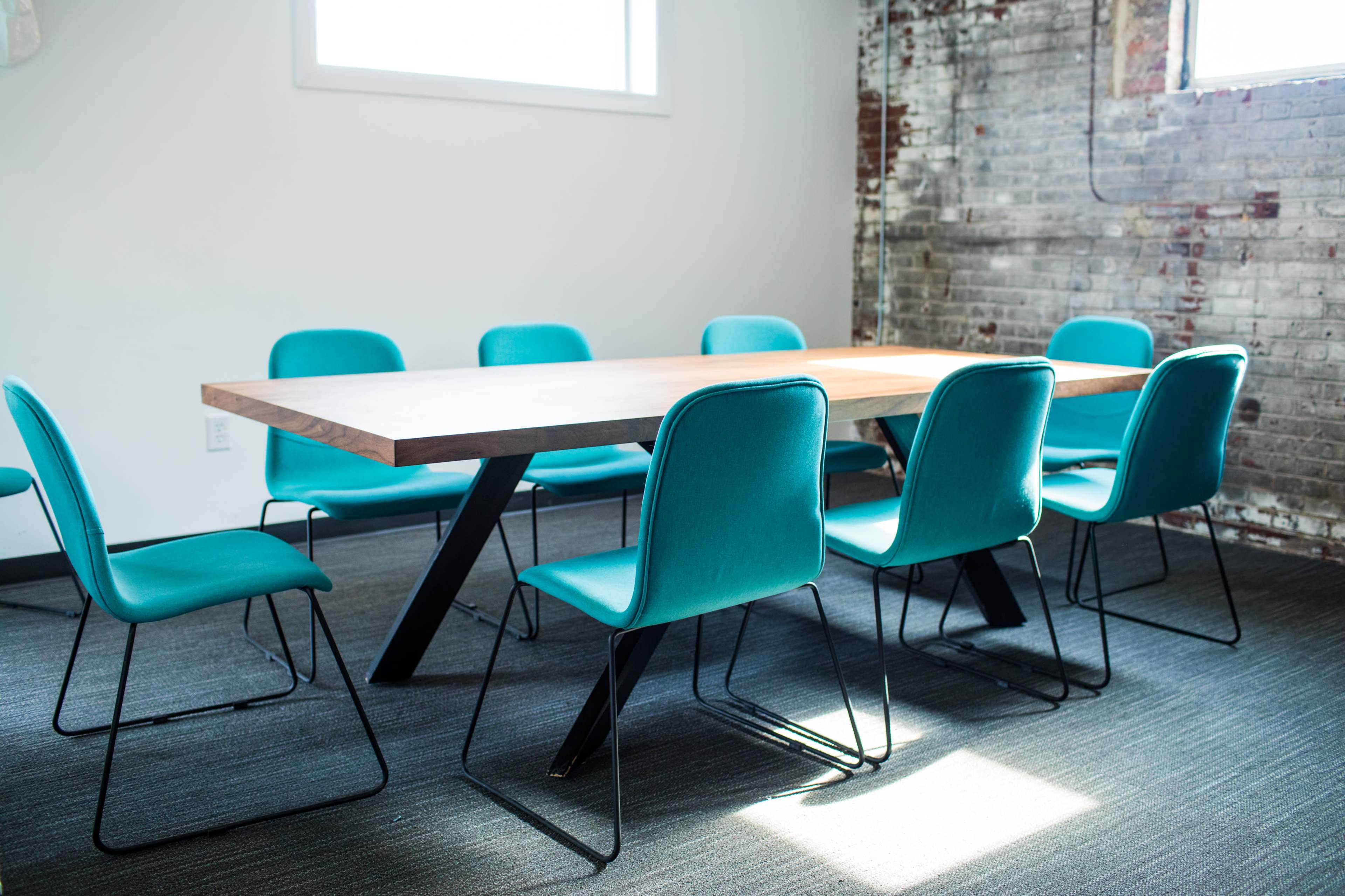 A spacious meeting room features a long wooden table surrounded by eight turquoise chairs against a brick wall.