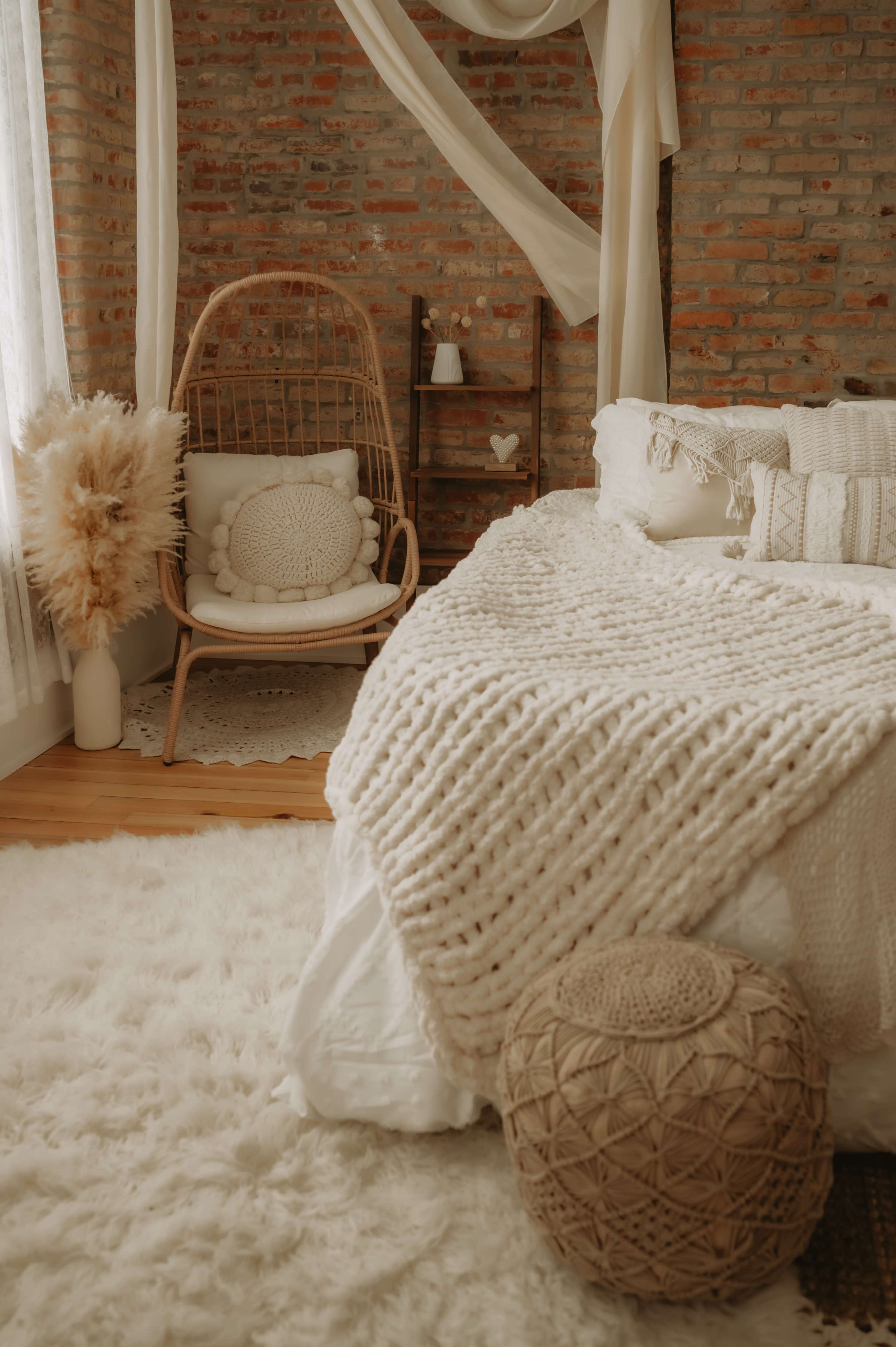 The image shows a cozy bedroom featuring a knitted blanket on the bed, a rattan chair in the corner, and a textured rug on the wooden floor.
