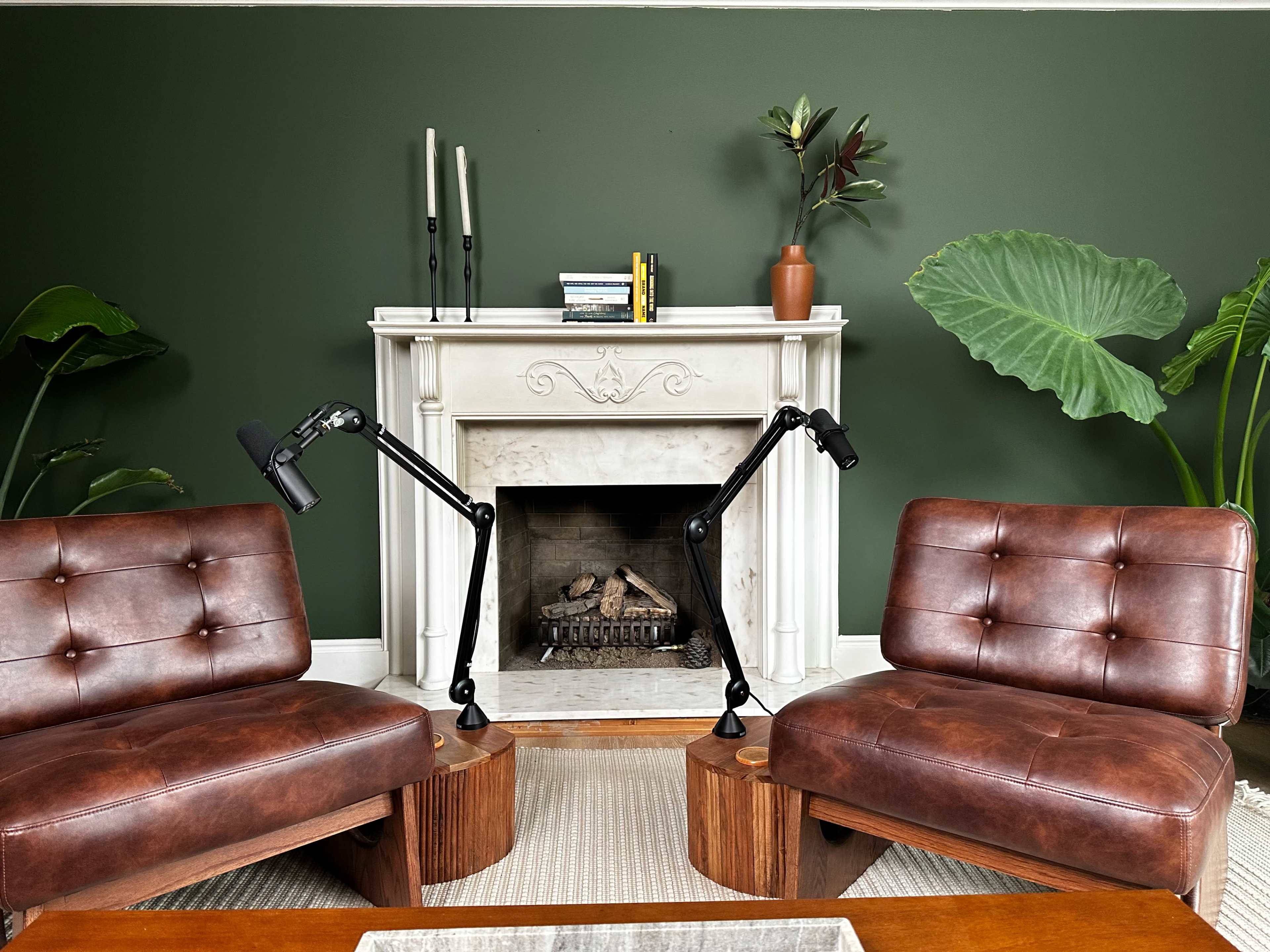 The image shows two brown leather chairs with wooden side tables facing a white marble fireplace on a green wall, surrounded by large indoor plants.