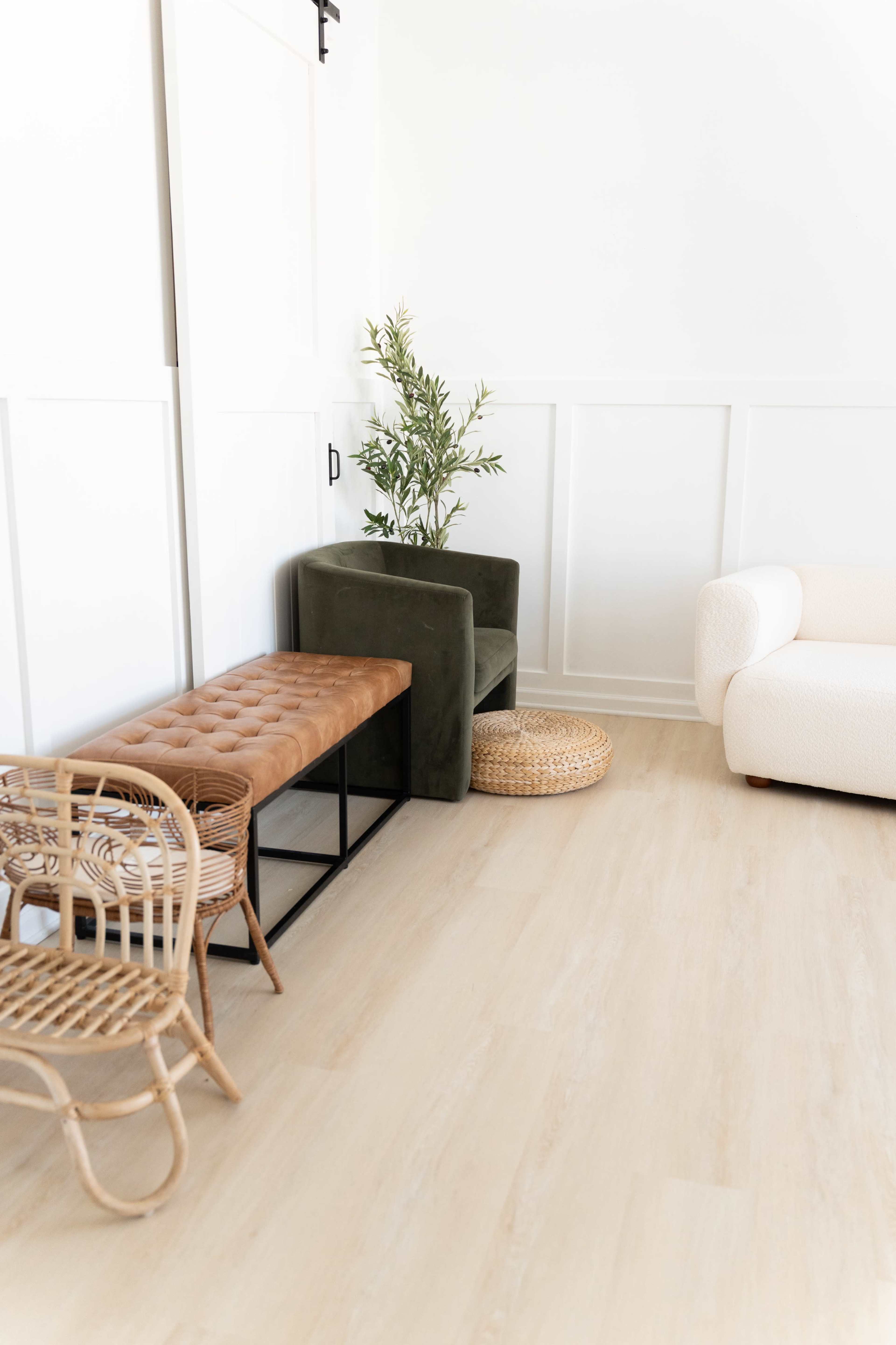 The image shows a modern living space featuring a green armchair, a tufted brown bench, a round woven basket, and a light-colored sofa against a white paneled wall.