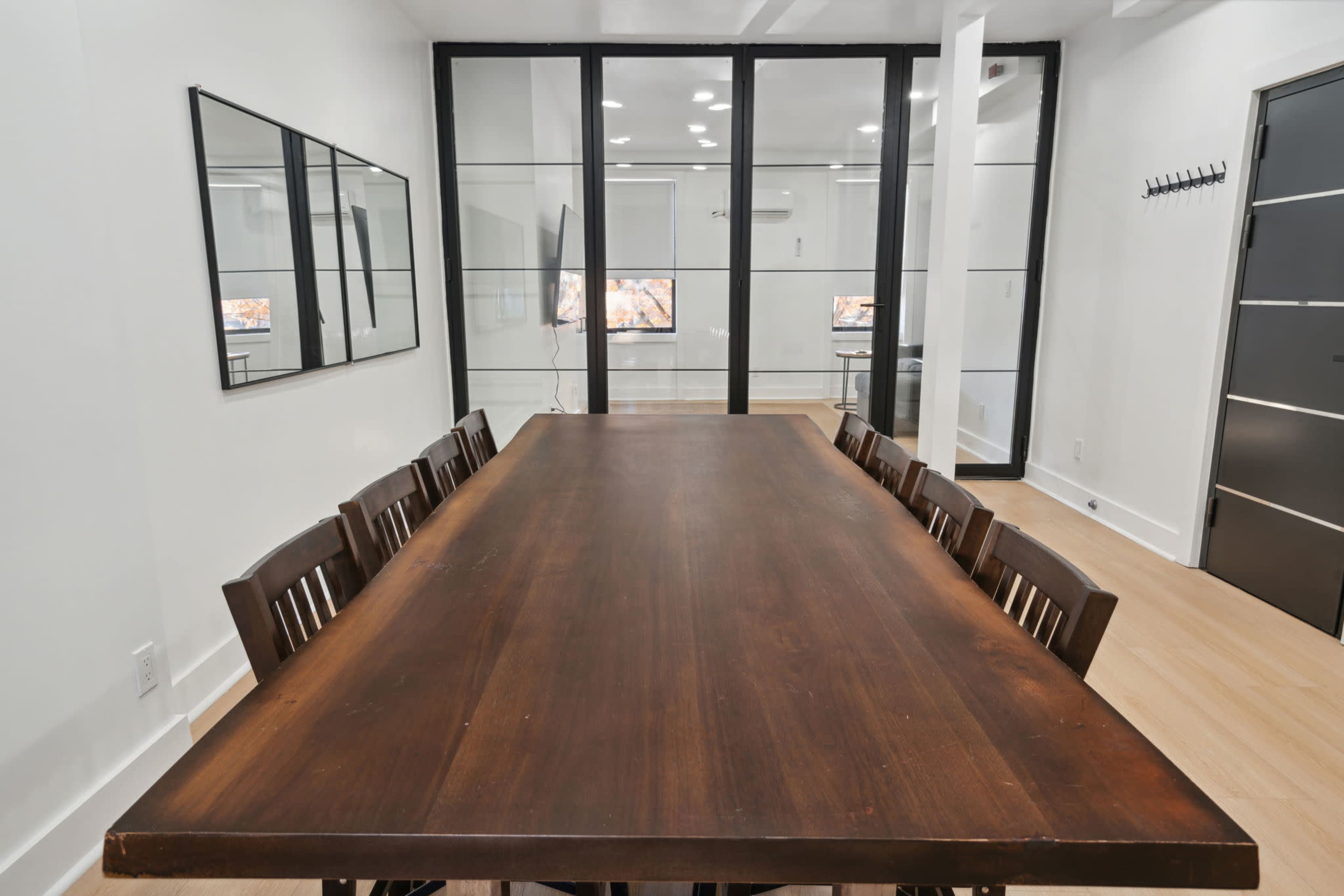 A long wooden table with eight chairs is positioned in a well-lit room separated by glass walls, featuring two screens visible in the background.