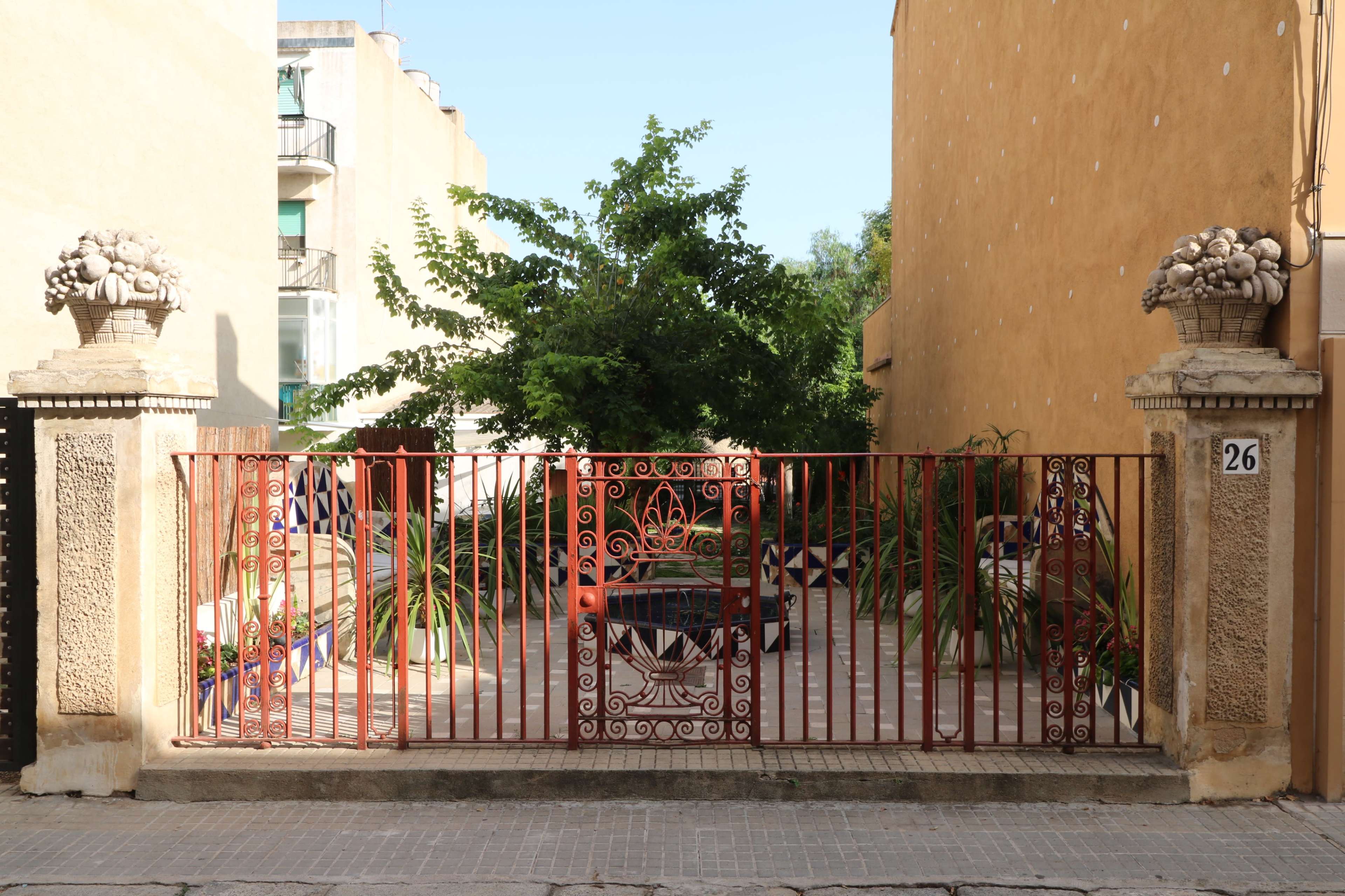 A decorative red metal gate leading to a garden area with potted plants, framed by stone columns topped with baskets.