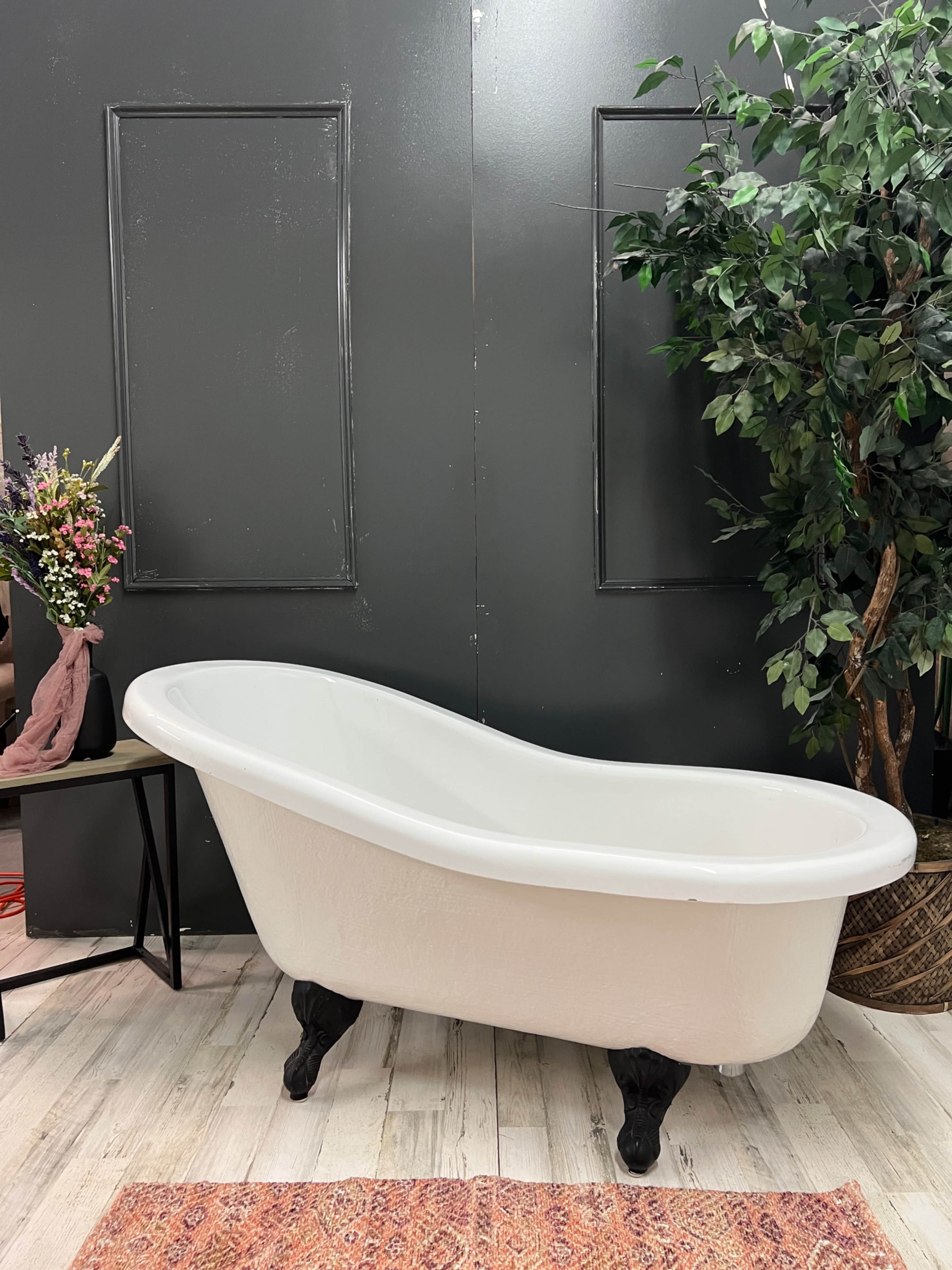 A white vintage-style bathtub on claw feet positioned against a dark wall, accompanied by a potted plant and a floral arrangement on a small table.