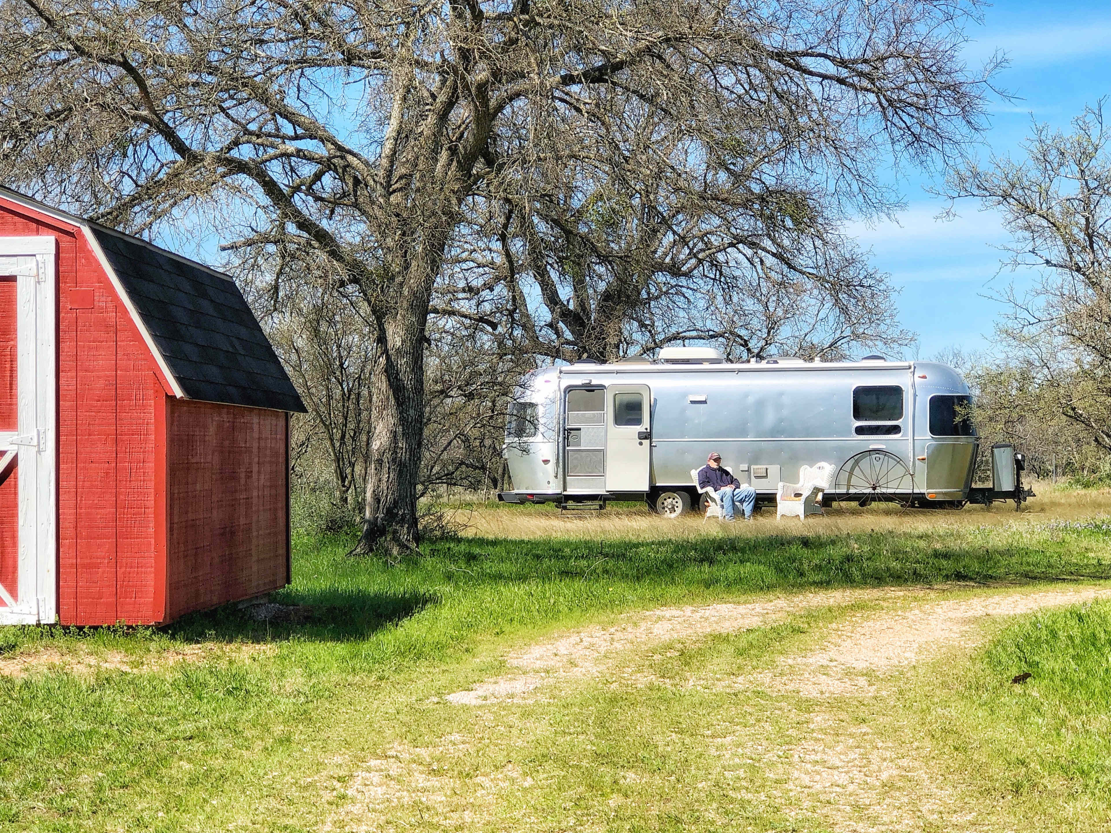 A silver Airstream trailer is parked under trees, beside a red shed, on a grassy path.