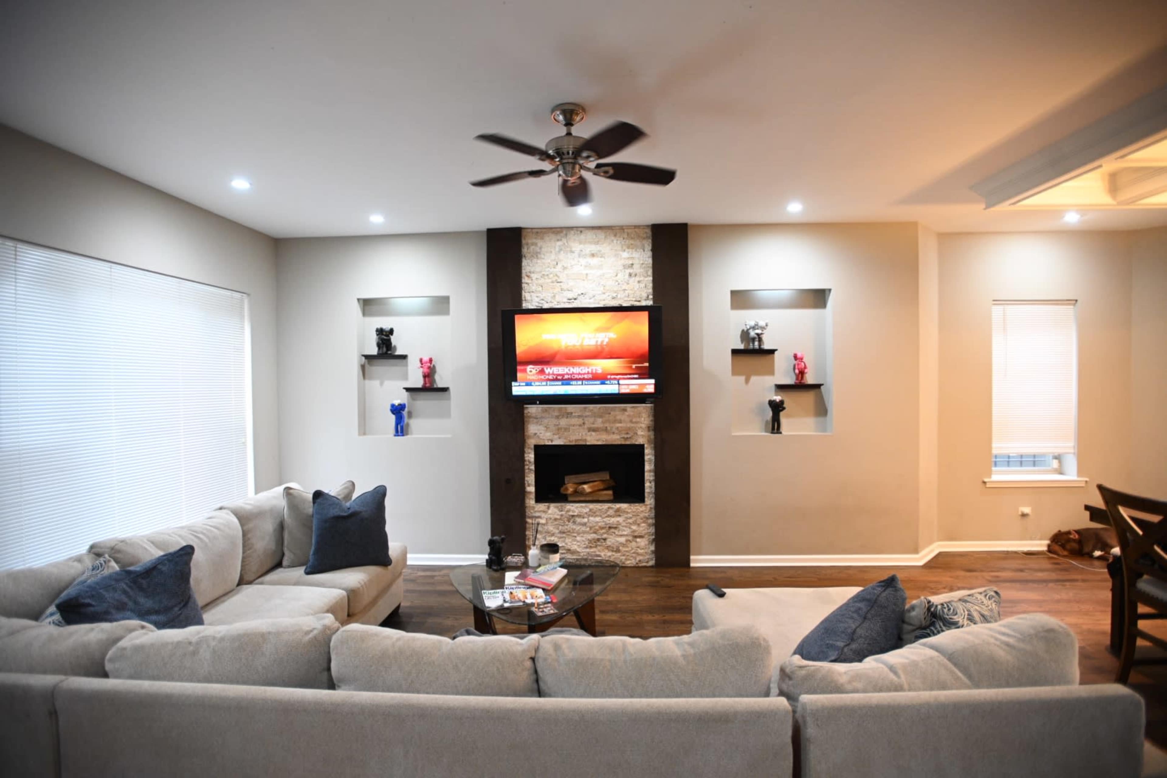 A spacious living room features a gray sectional sofa, a stone fireplace with a mounted TV above it, and decorative shelves with contrasting-colored figurines.
