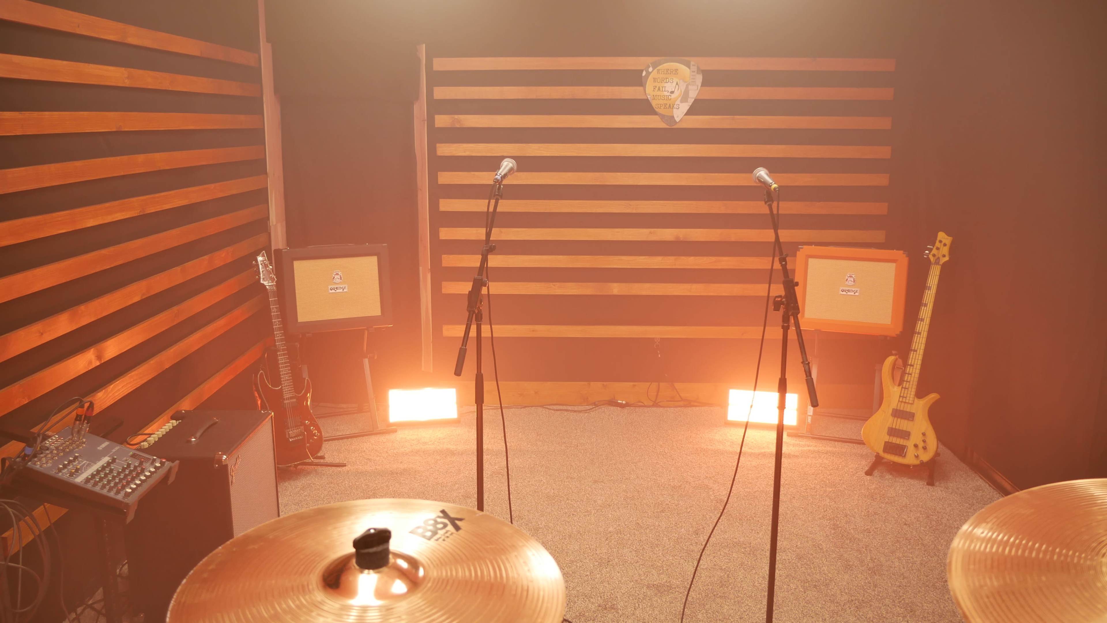 A music rehearsal space features two microphones on stands, two amplifiers, and various instruments amidst warm lighting and wooden wall slats.