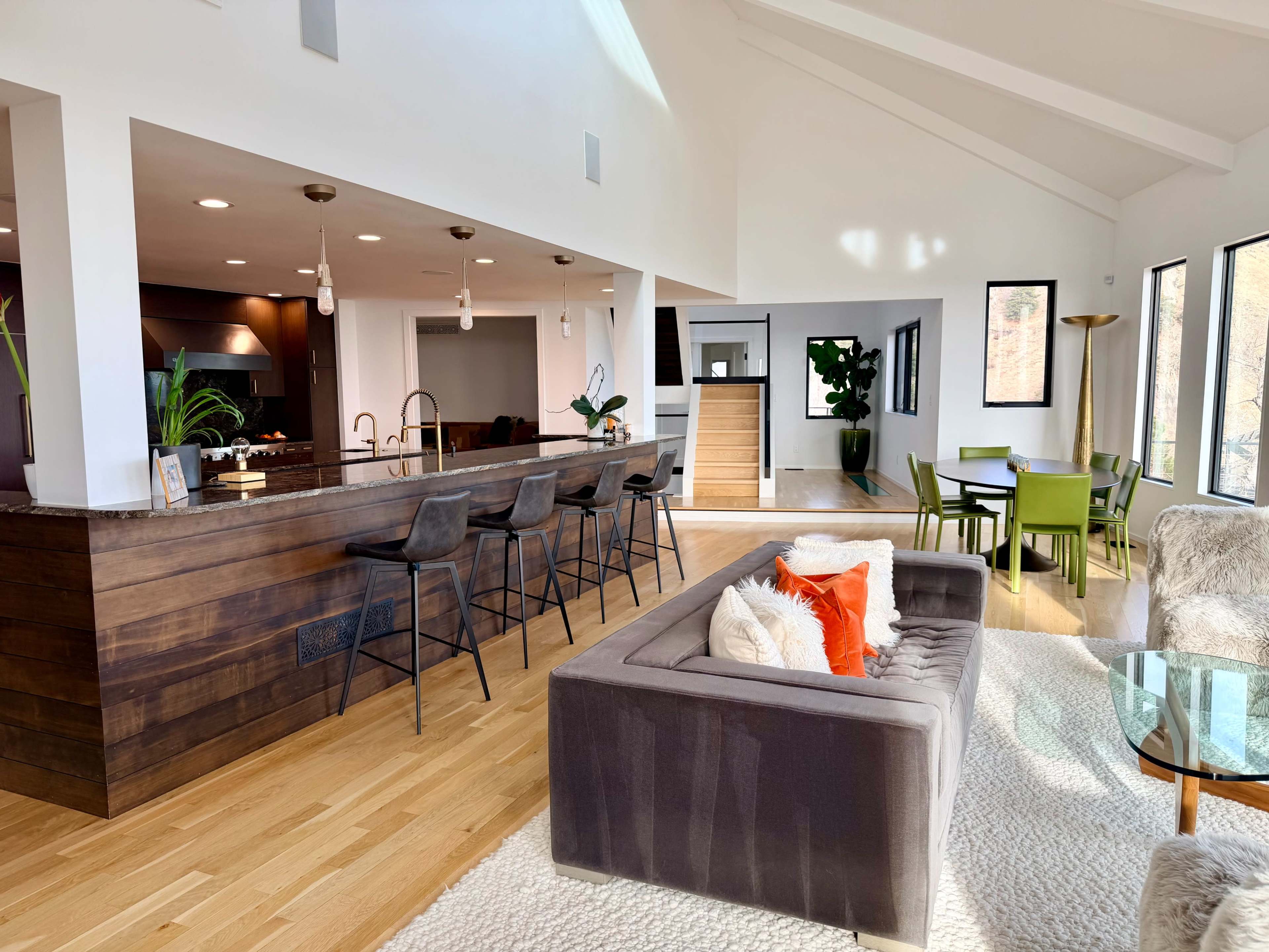 The image shows a modern open-concept living area featuring a dark sofa, a kitchen island with bar stools, and a dining area with green chairs, illuminated by natural light from large windows.