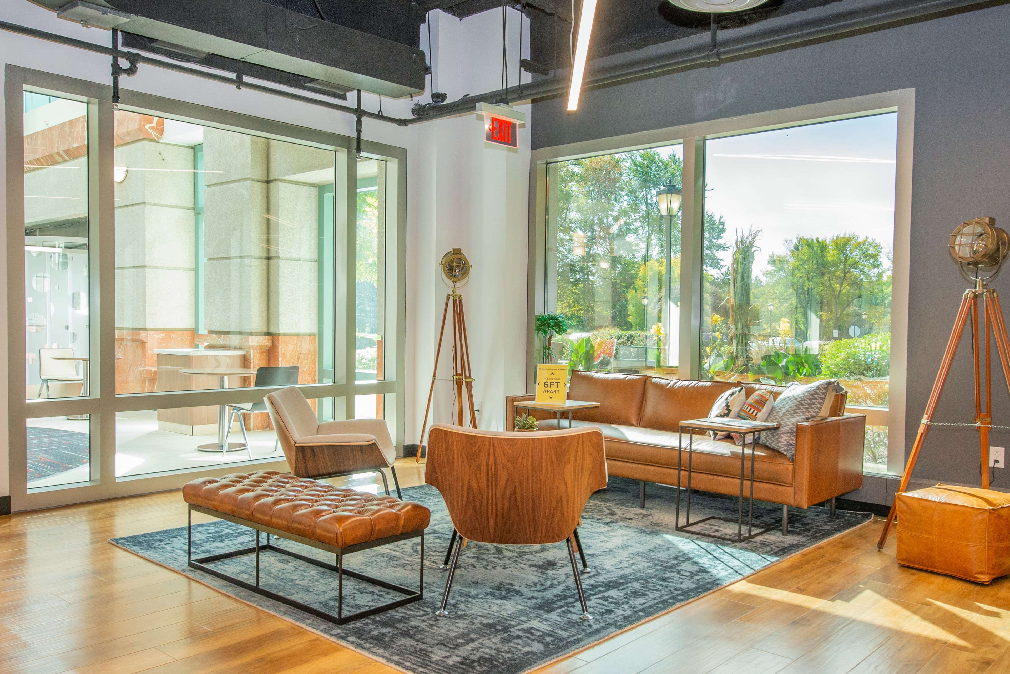 The image shows a modern lounge area with a brown leather sofa, an armchair, a footrest, and large windows overlooking a garden.