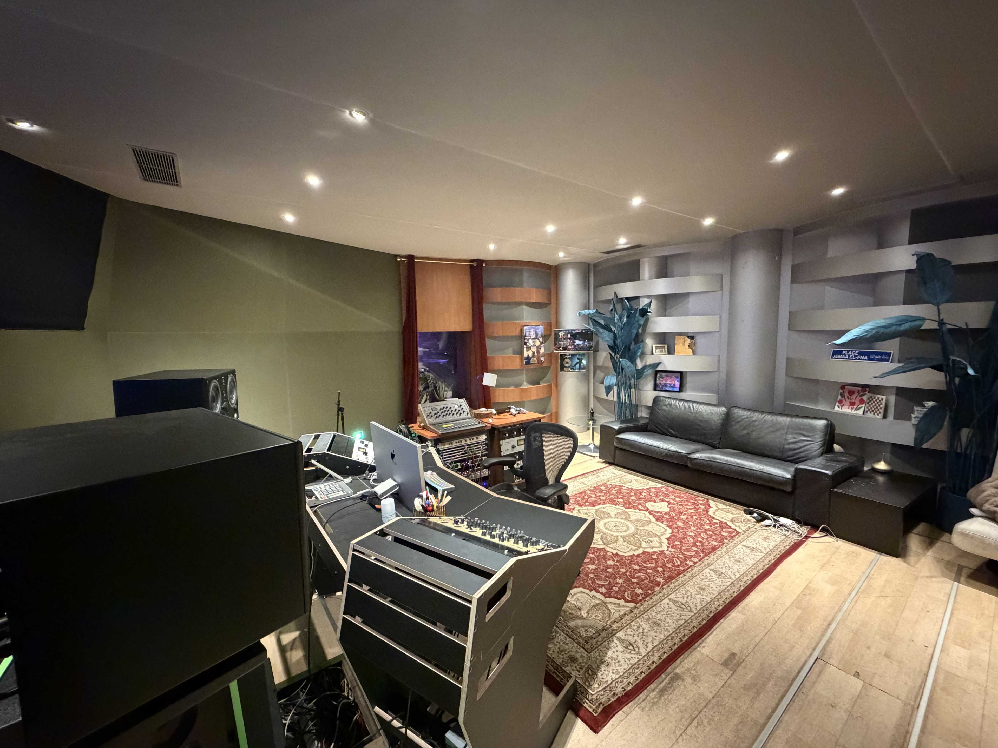 The image shows a recording studio with sound equipment, a black couch, a decorative rug, and shelves displaying various items.
