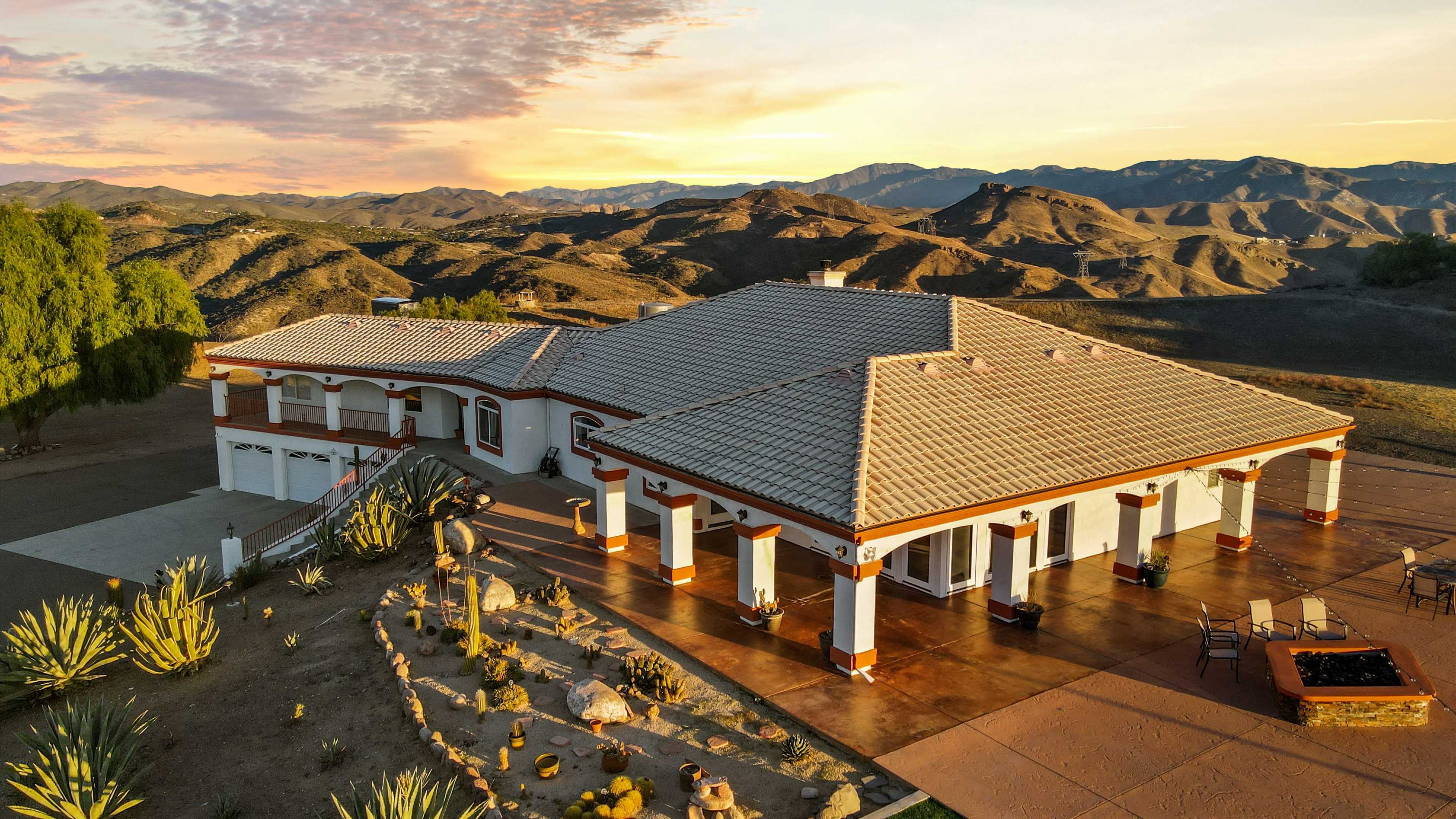 A single-story house with a tiled roof is situated in a mountainous landscape, surrounded by desert plants and an expansive patio.