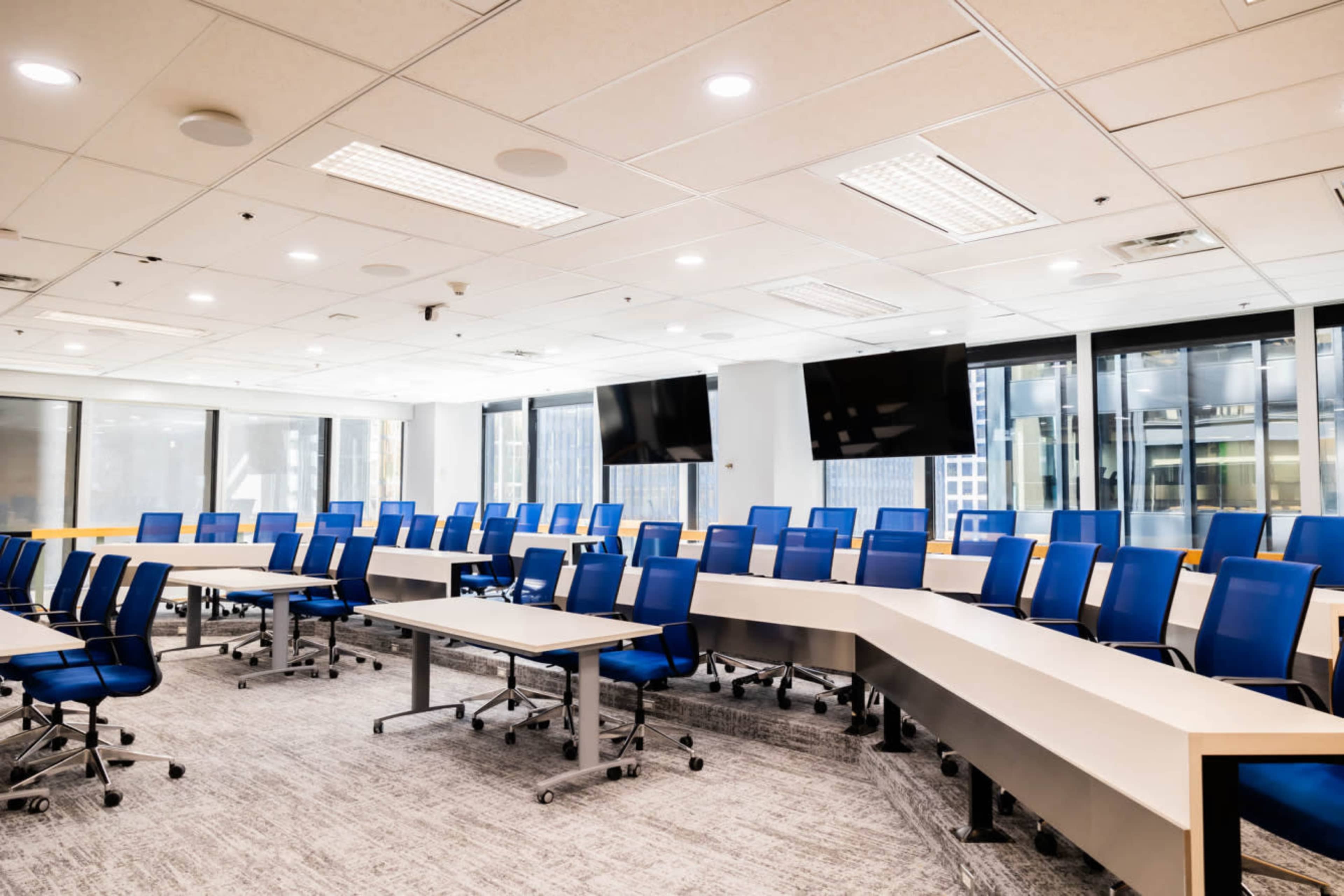 A modern conference room features multiple rows of blue chairs and tables arranged in a curved layout, with large windows and two mounted screens.