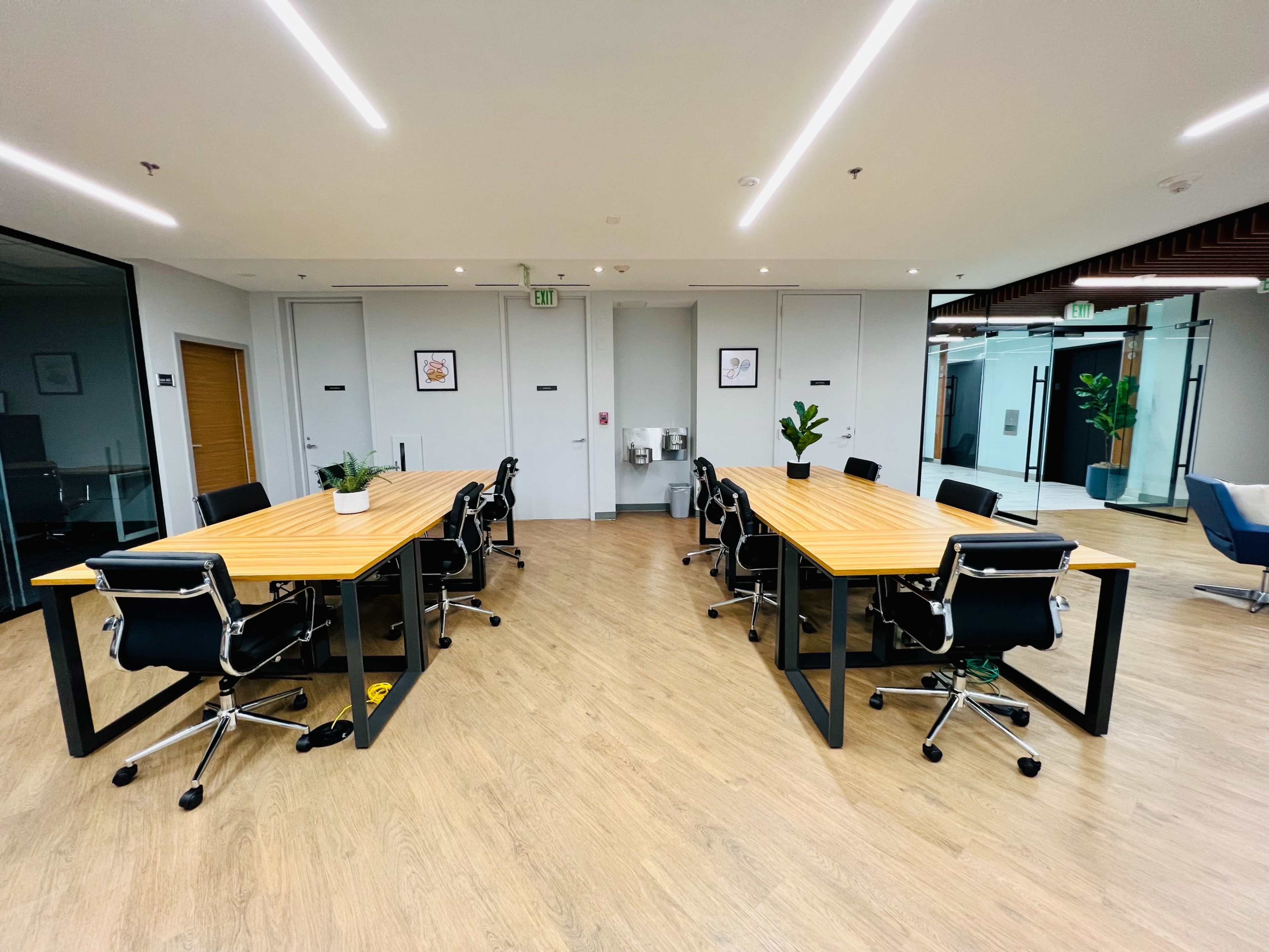 A modern office space features two large conference tables surrounded by black ergonomic chairs, with plants and artwork on the walls.