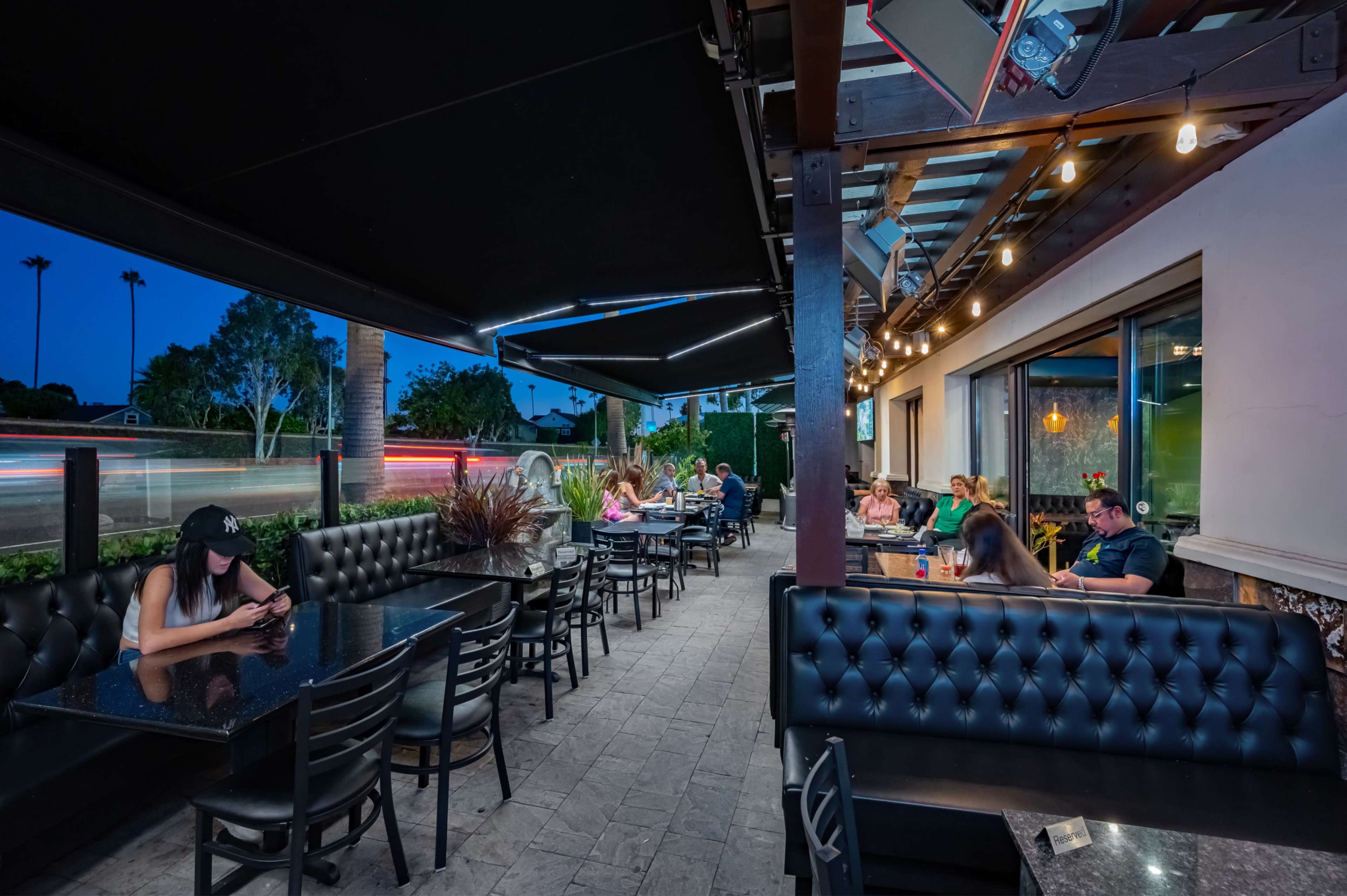 The image shows an outdoor restaurant patio with several tables and patrons seated, illuminated by string lights against a twilight backdrop.