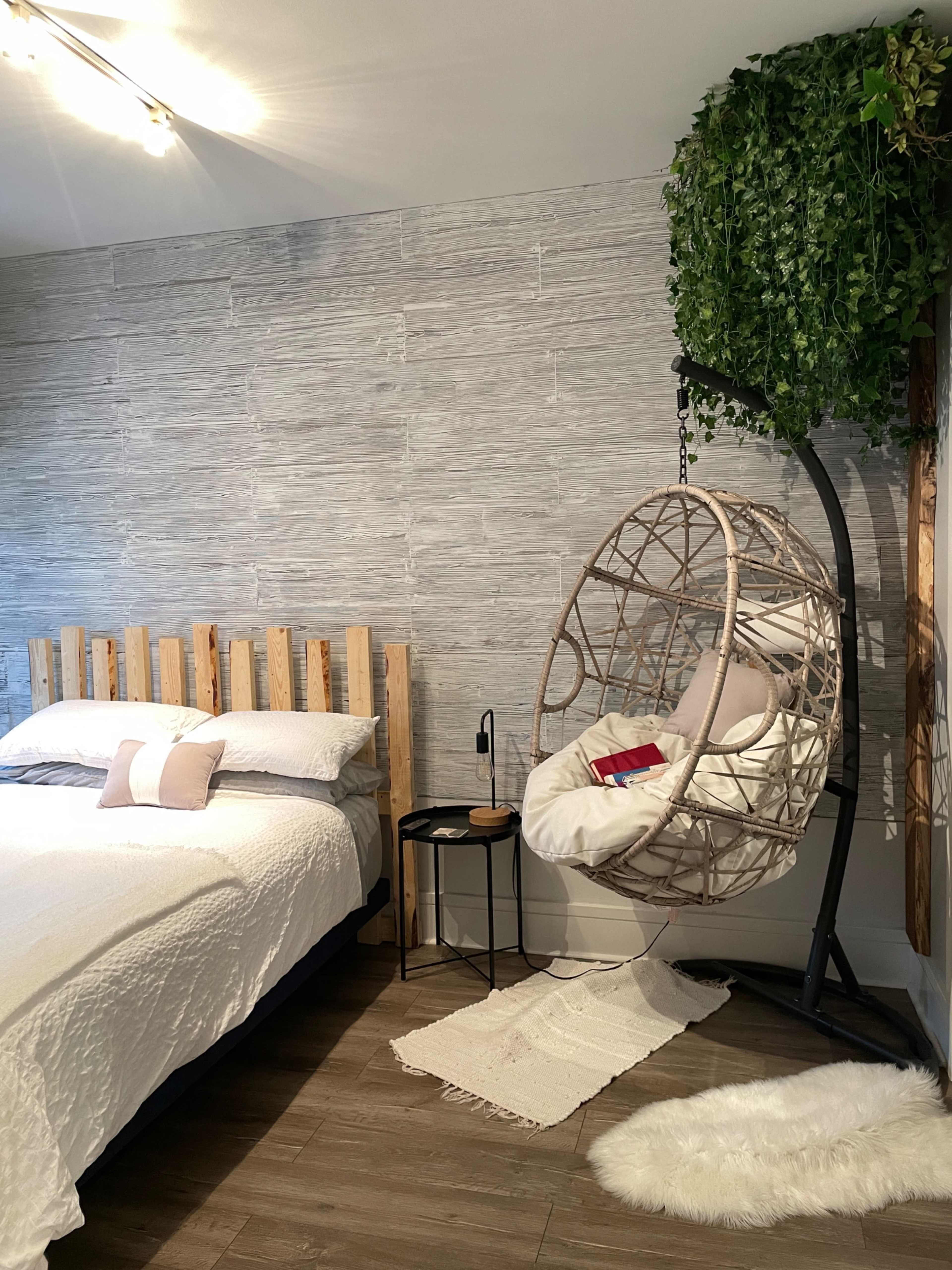A bedroom features a wooden slatted bed, a hanging swing chair, and a side table against a textured gray wall.