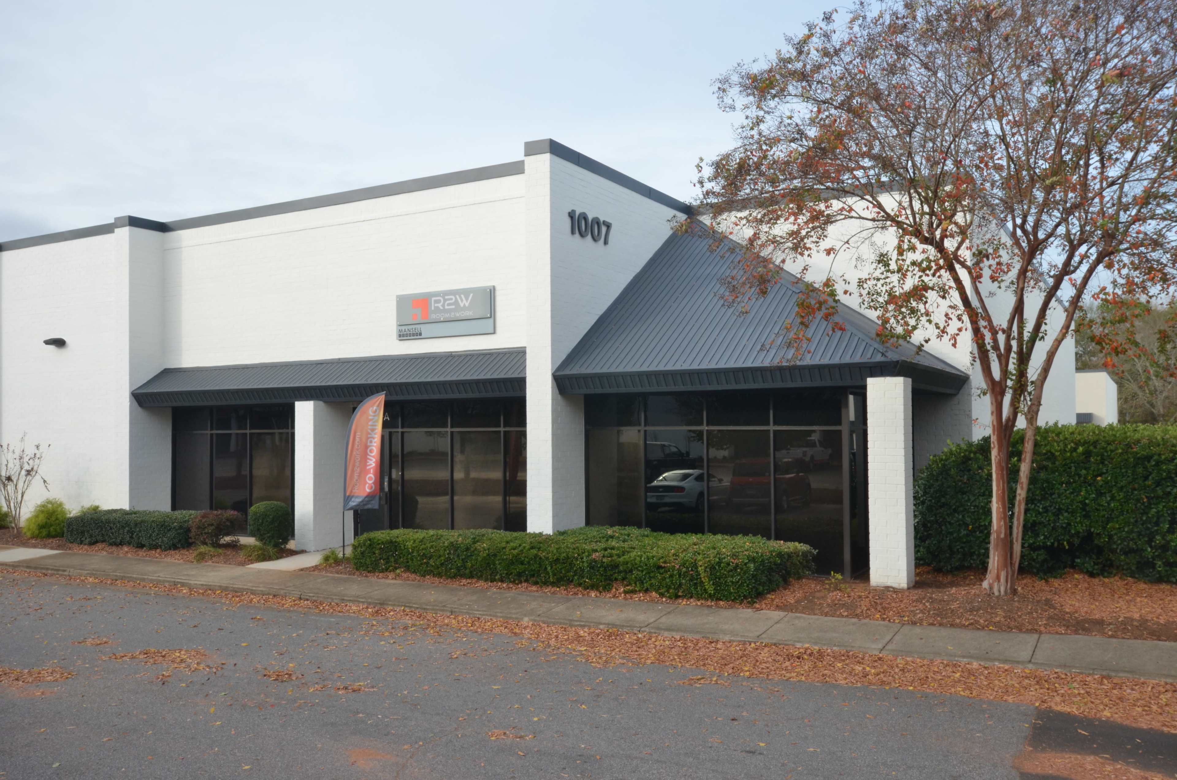 A white commercial building with a dark gray metal roof, featuring large windows and a sign that reads "R2W," located on a clean street.