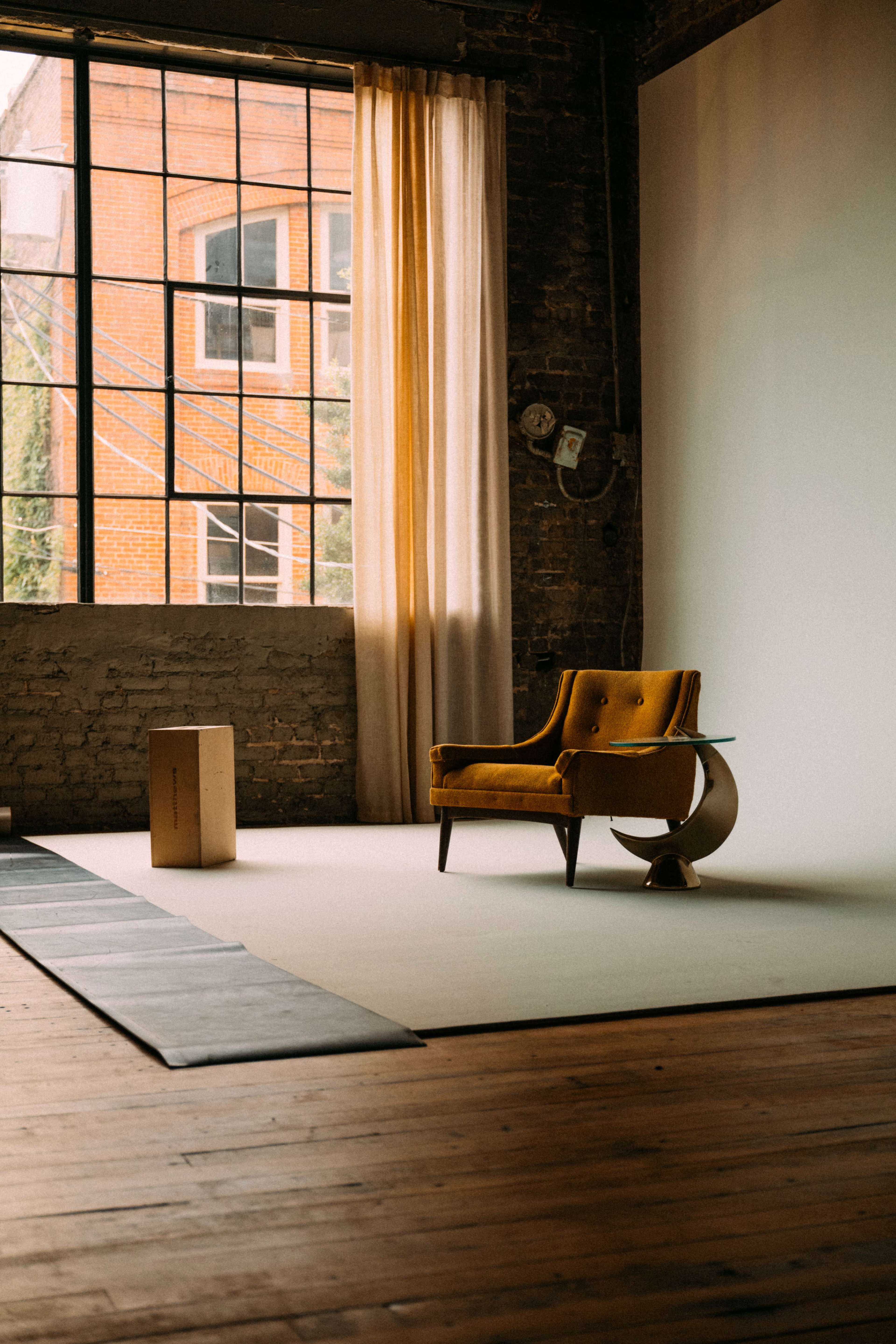 A yellow chair is positioned next to a wooden box on a light-colored floor in a room with large windows allowing natural light to enter.