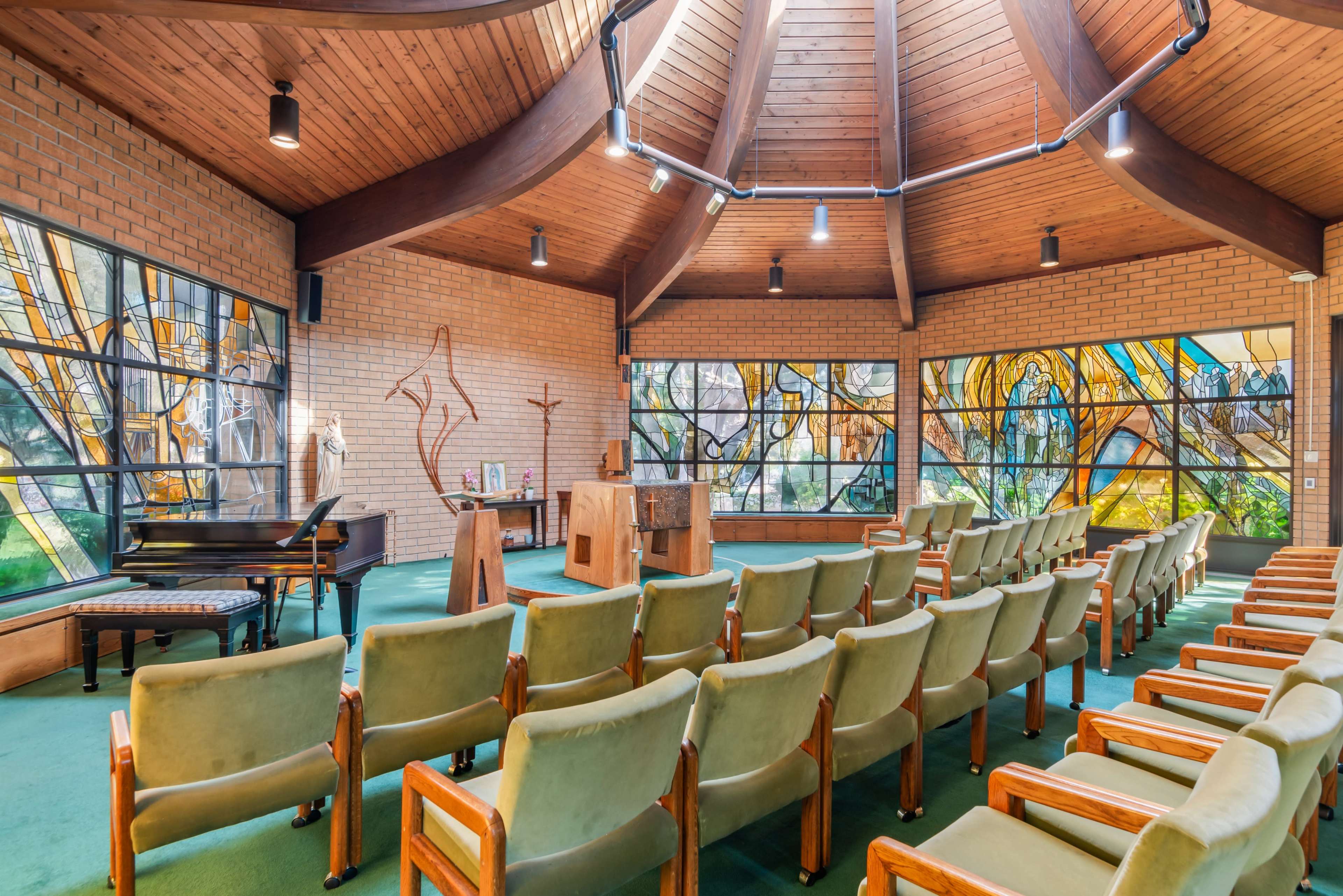 The interior of a chapel features rows of green upholstered chairs facing a wooden altar and colorful stained glass windows.