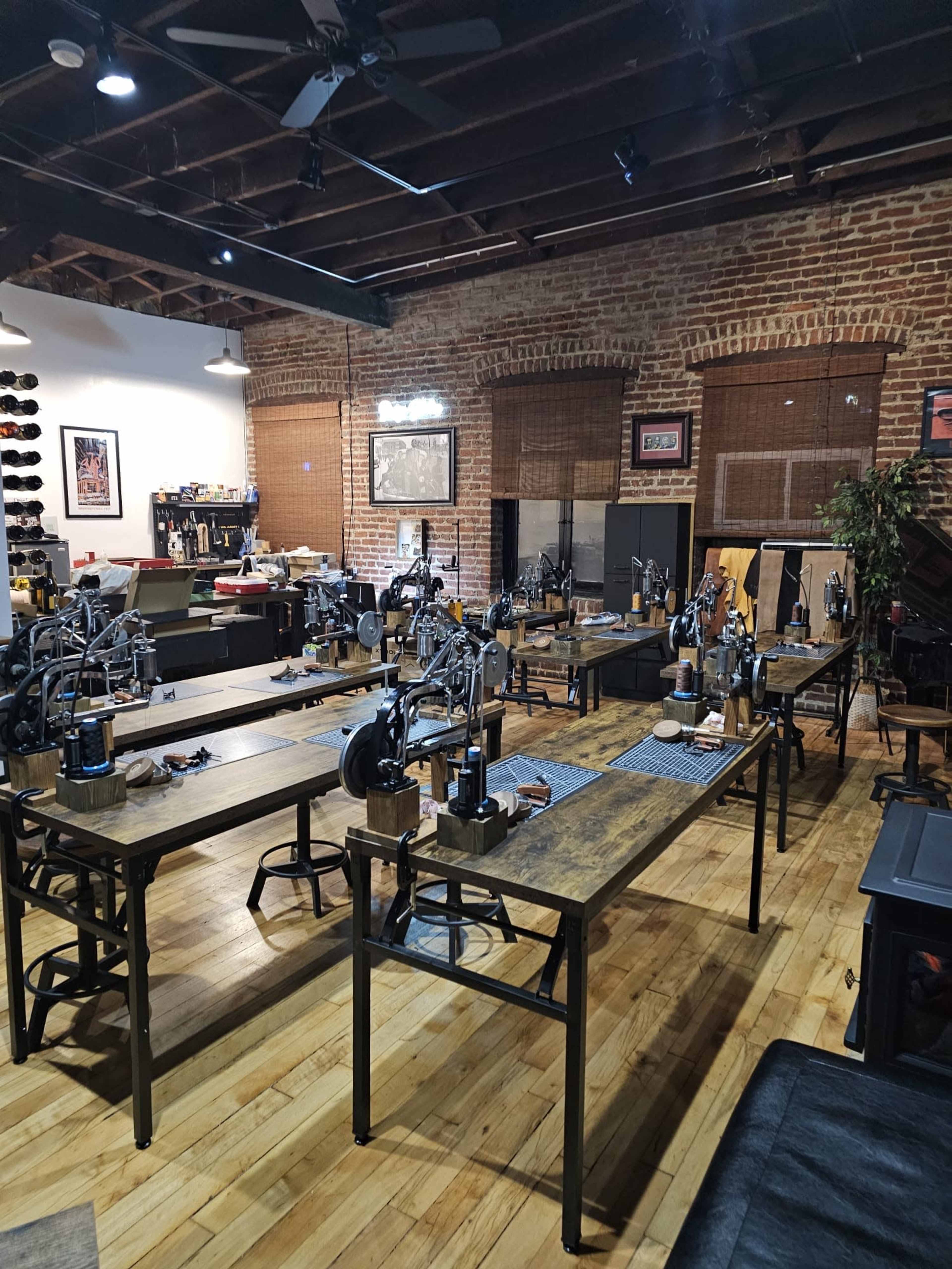 The image depicts a spacious workshop featuring multiple work tables equipped with sewing machines, hardwood flooring, exposed brick walls, and industrial-style lighting.