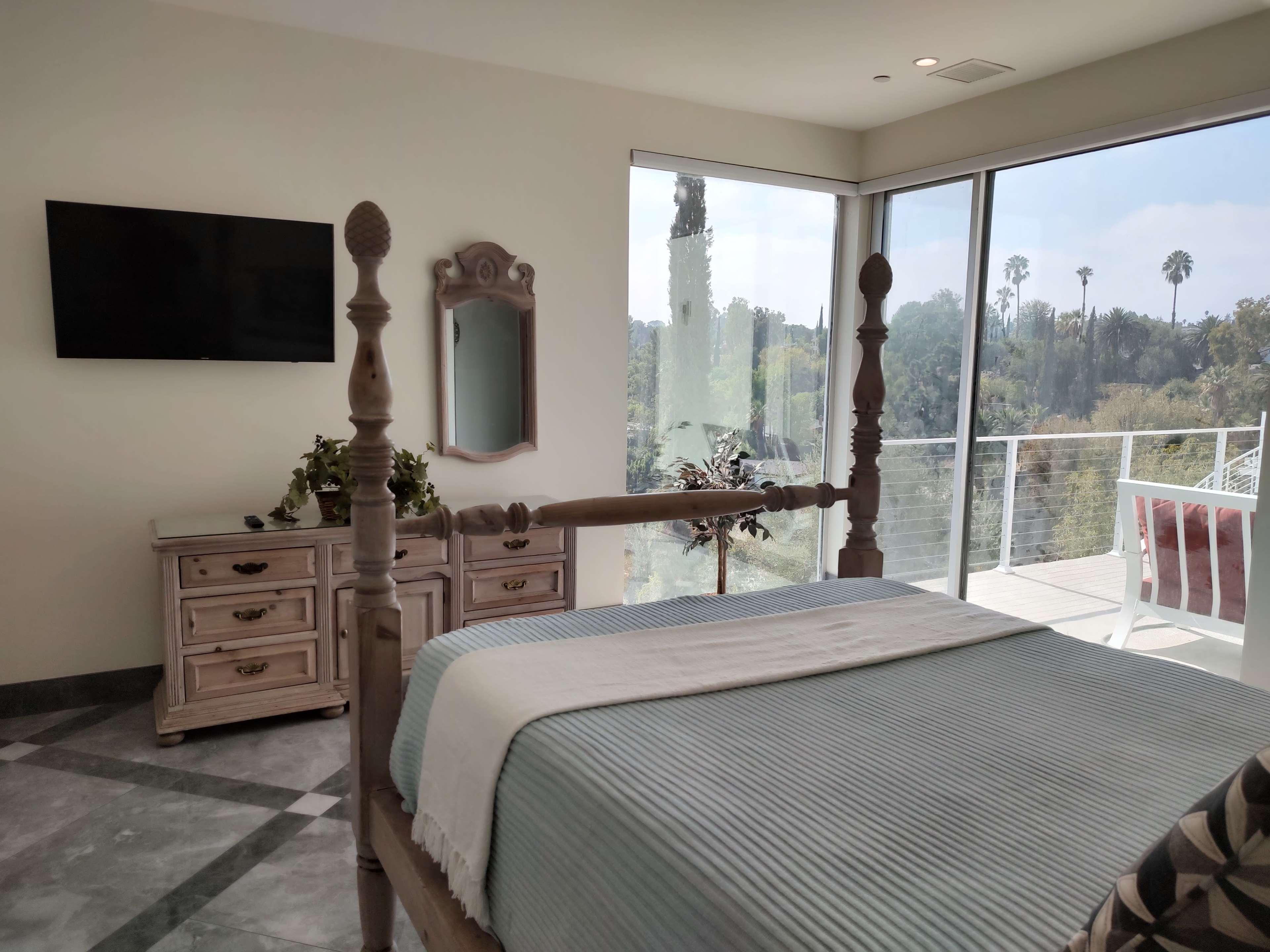 A modern bedroom with a wooden bed, a dresser, and large windows overlooking a landscaped area with palm trees.