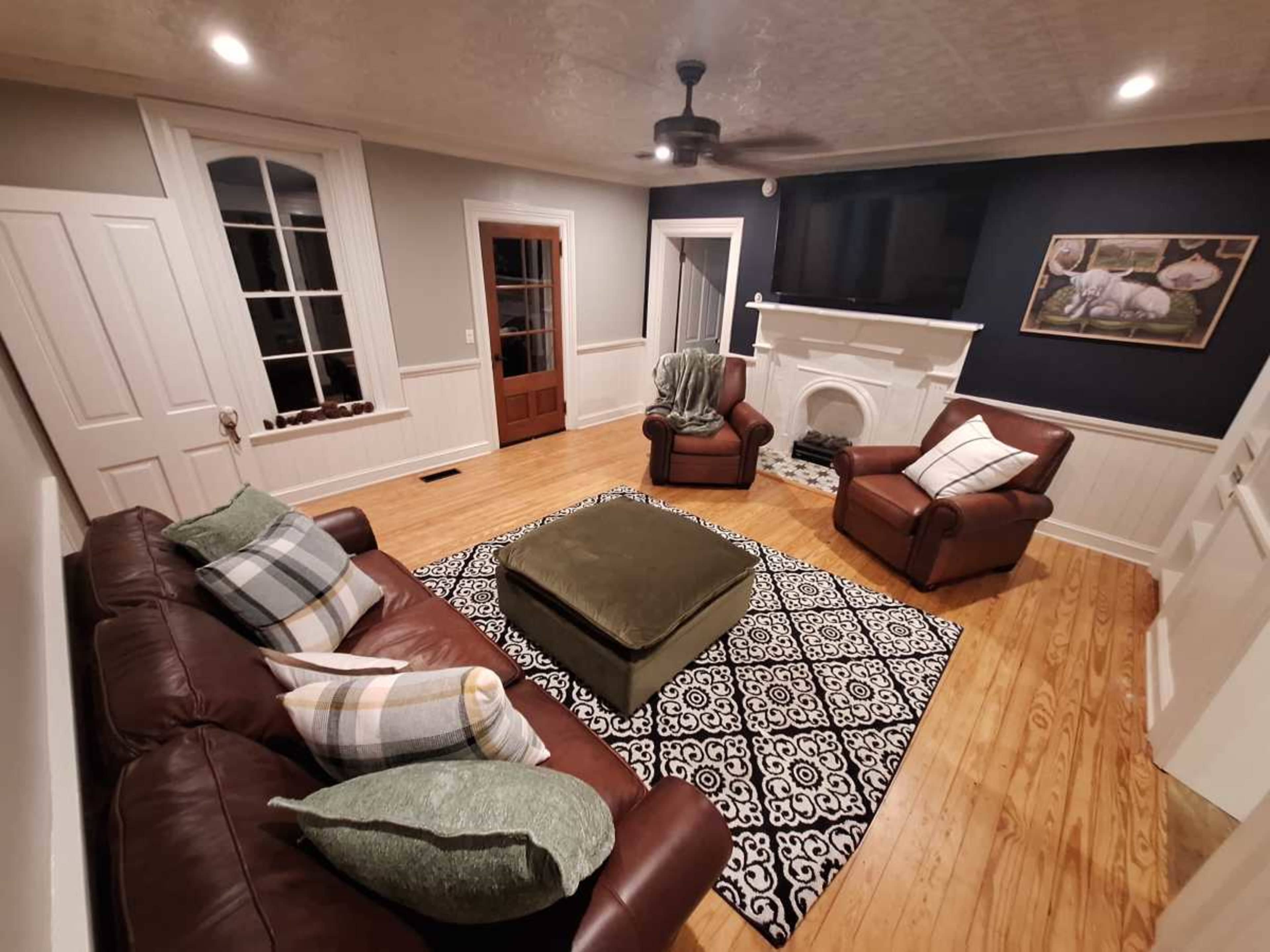 The living room features a leather sofa and armchairs arranged around a patterned area rug, with a fireplace and a dark accent wall.