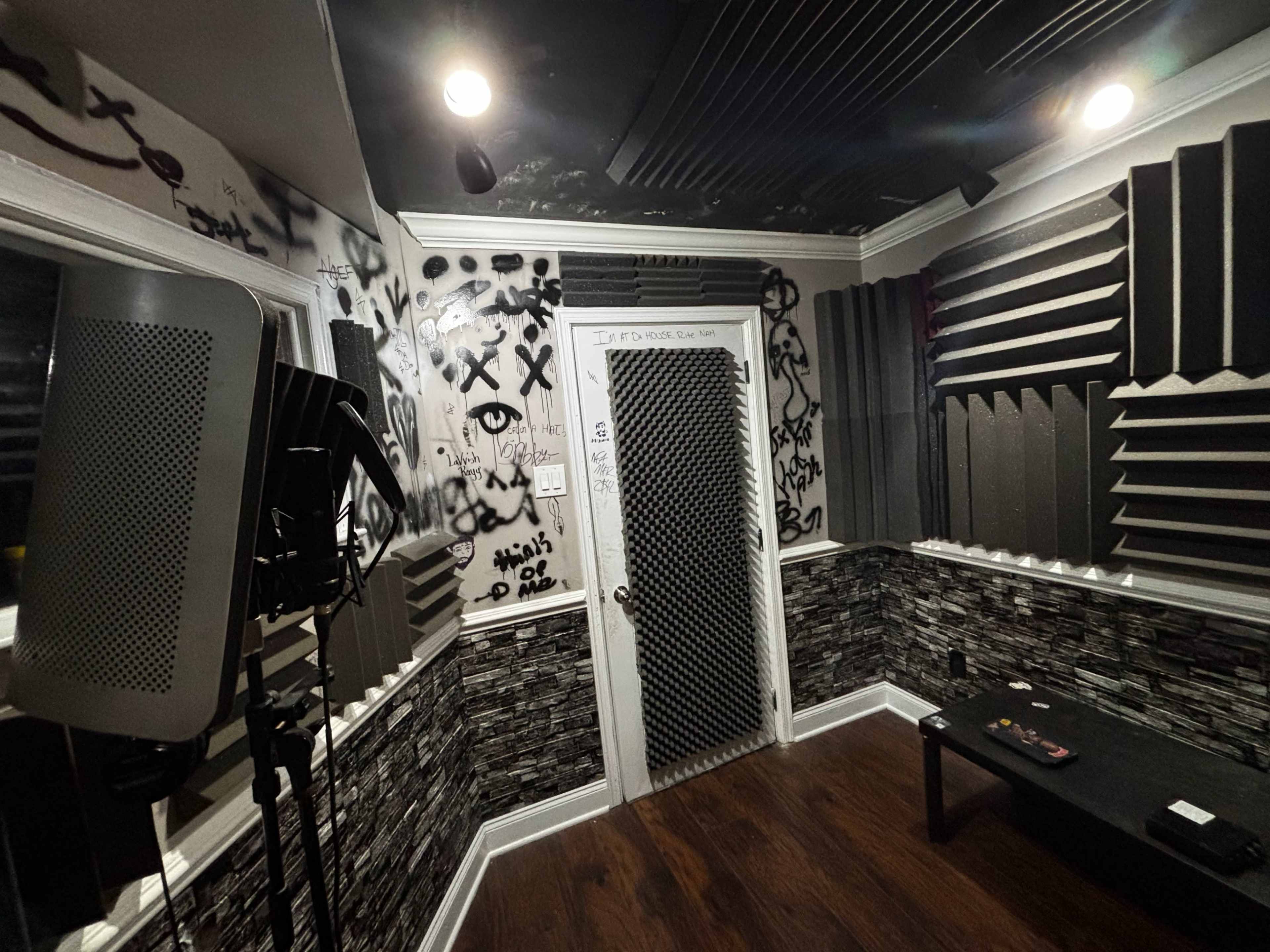 The image shows a small recording studio with soundproofing panels on the walls, graffiti on the walls, and a microphone setup in one corner.