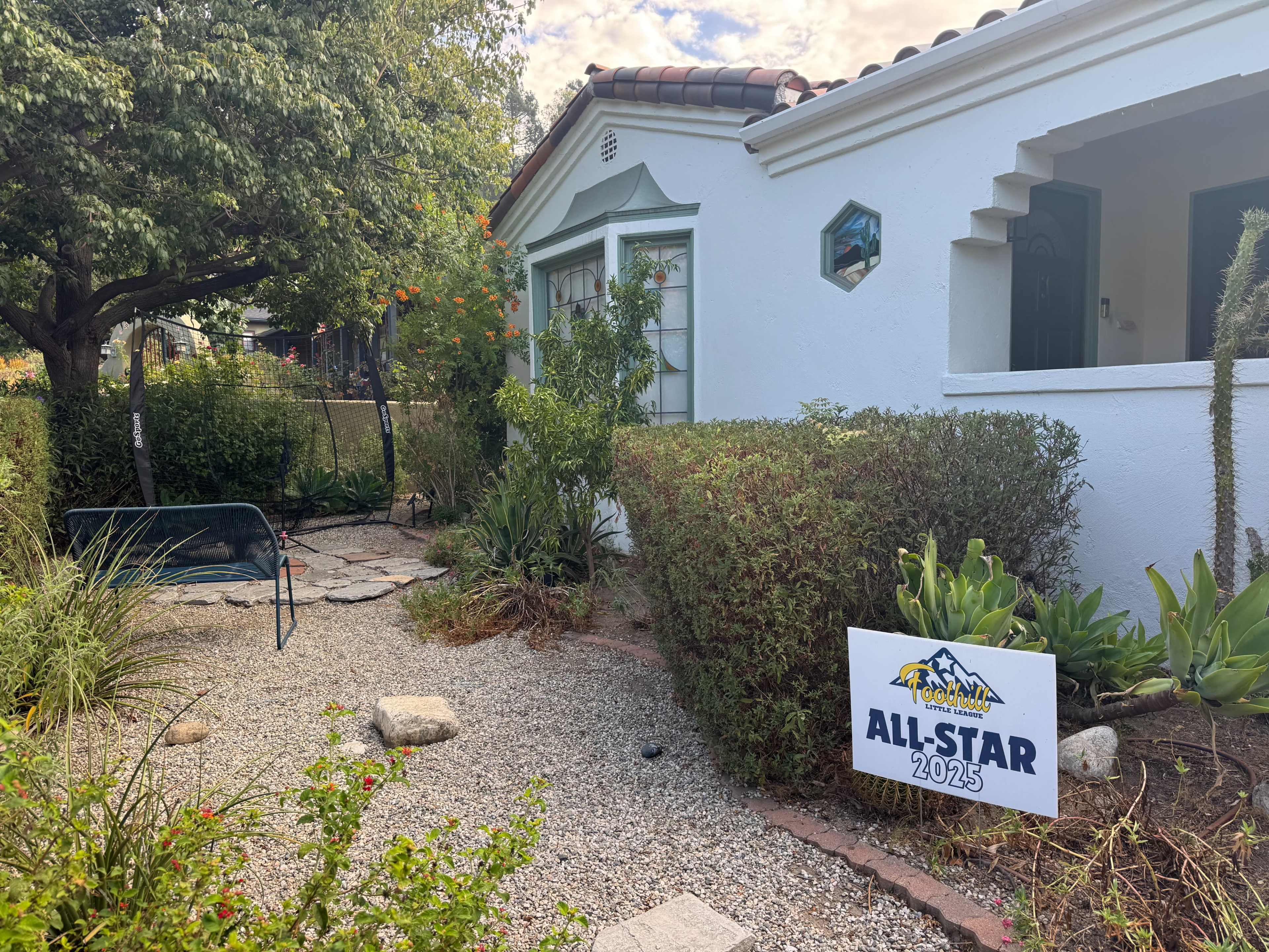 The image shows a residential garden with a white house facade, featuring a grassy area, various plants, and a sign that reads "ALL-STAR 2025."