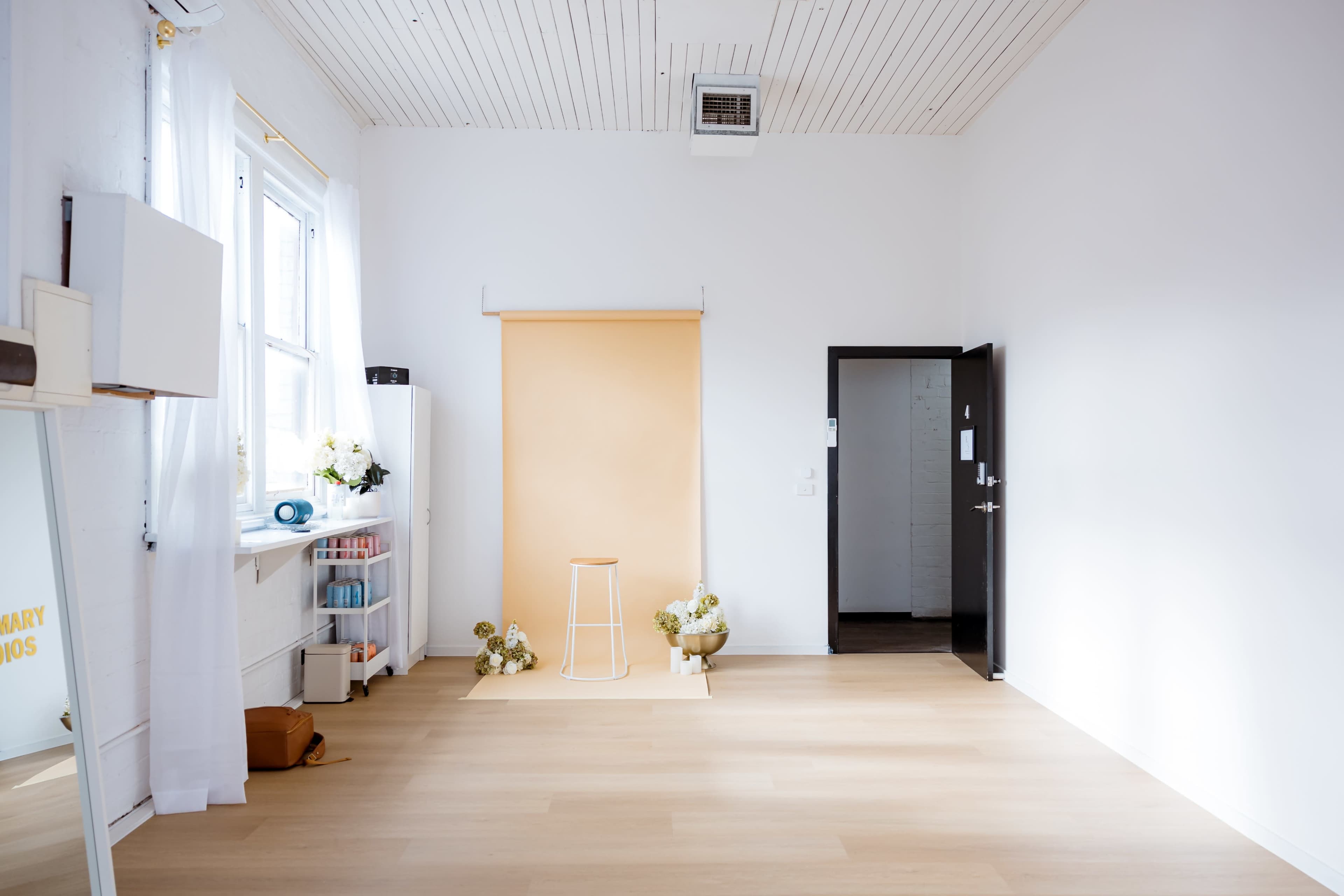 The image shows a bright, minimalistic room featuring a light wood floor, a peach backdrop set up for photography, a white stool, and decorative flowers placed near the backdrop.