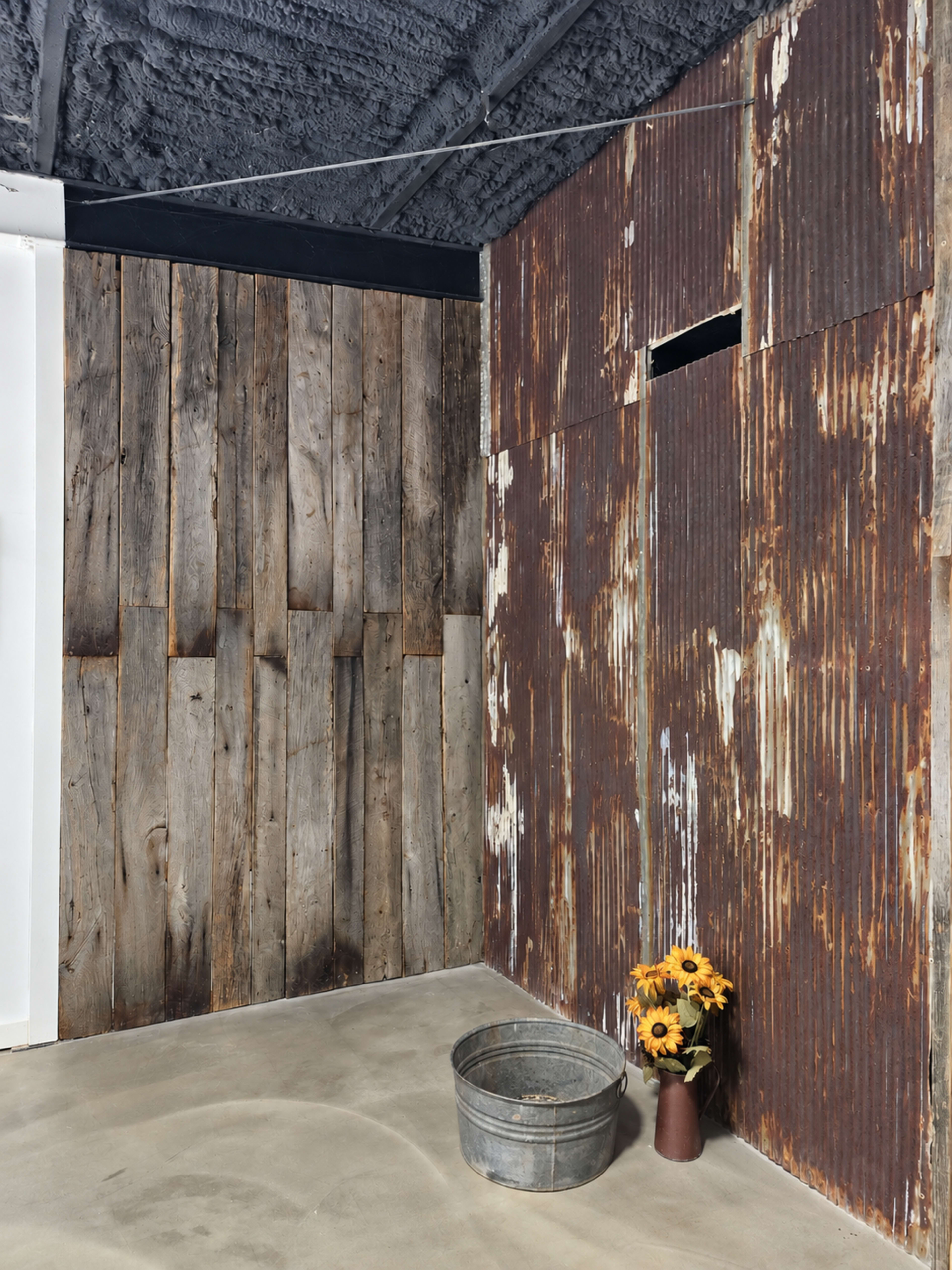 The image shows a corner of a room featuring a wooden wall and a rusted metal wall, with a metal bucket and a vase of sunflowers on the floor.