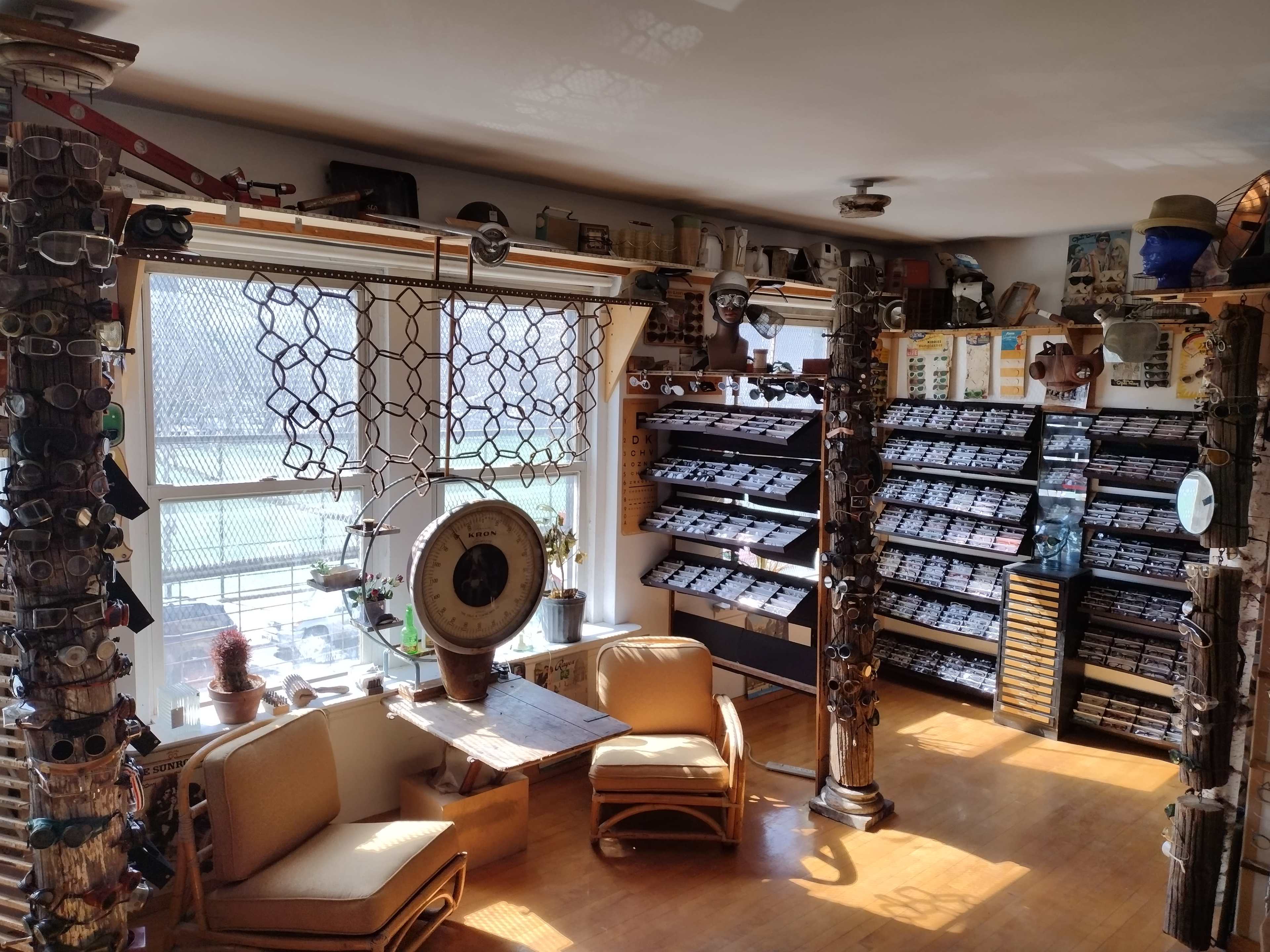 The image shows a well-organized room filled with various eyeglasses displayed on racks, with a large window providing natural light and two chairs positioned around a small table.