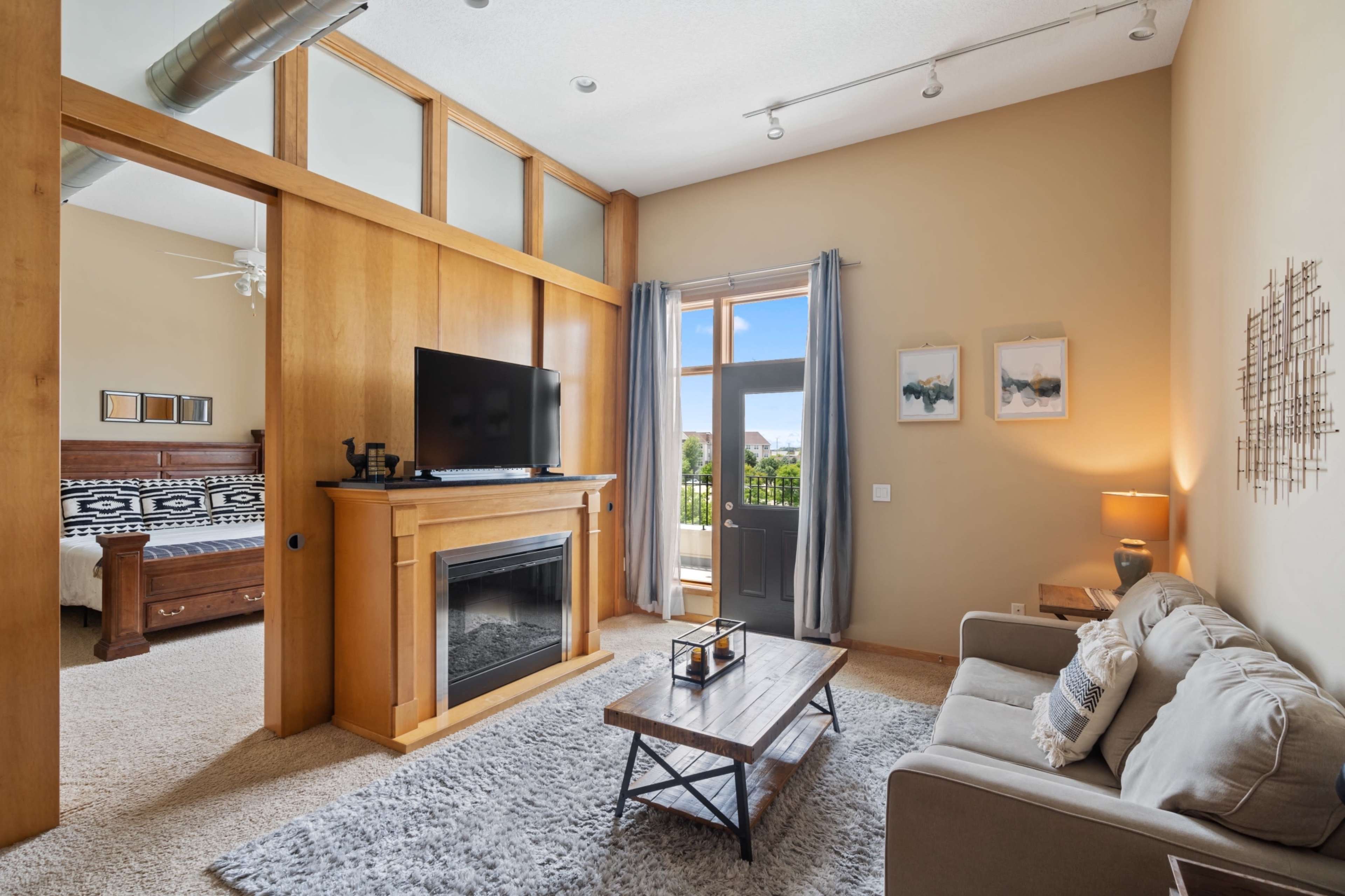 A cozy living room with a television mounted on a wooden mantel, a gray sofa, and large windows that provide natural light.