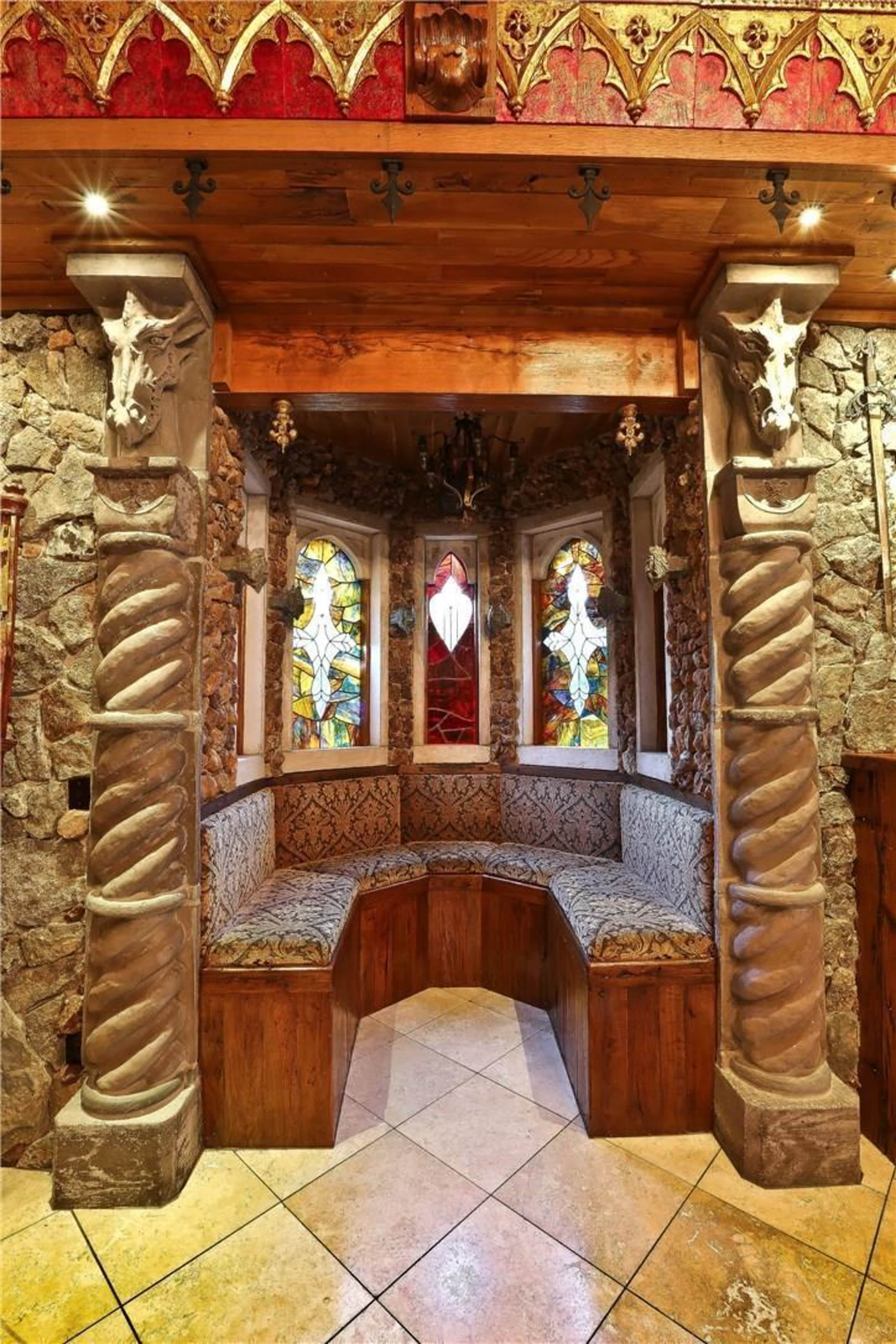 A nook with stone walls, carved pillar details, and stained glass windows, featuring a curved wooden bench with patterned upholstery.