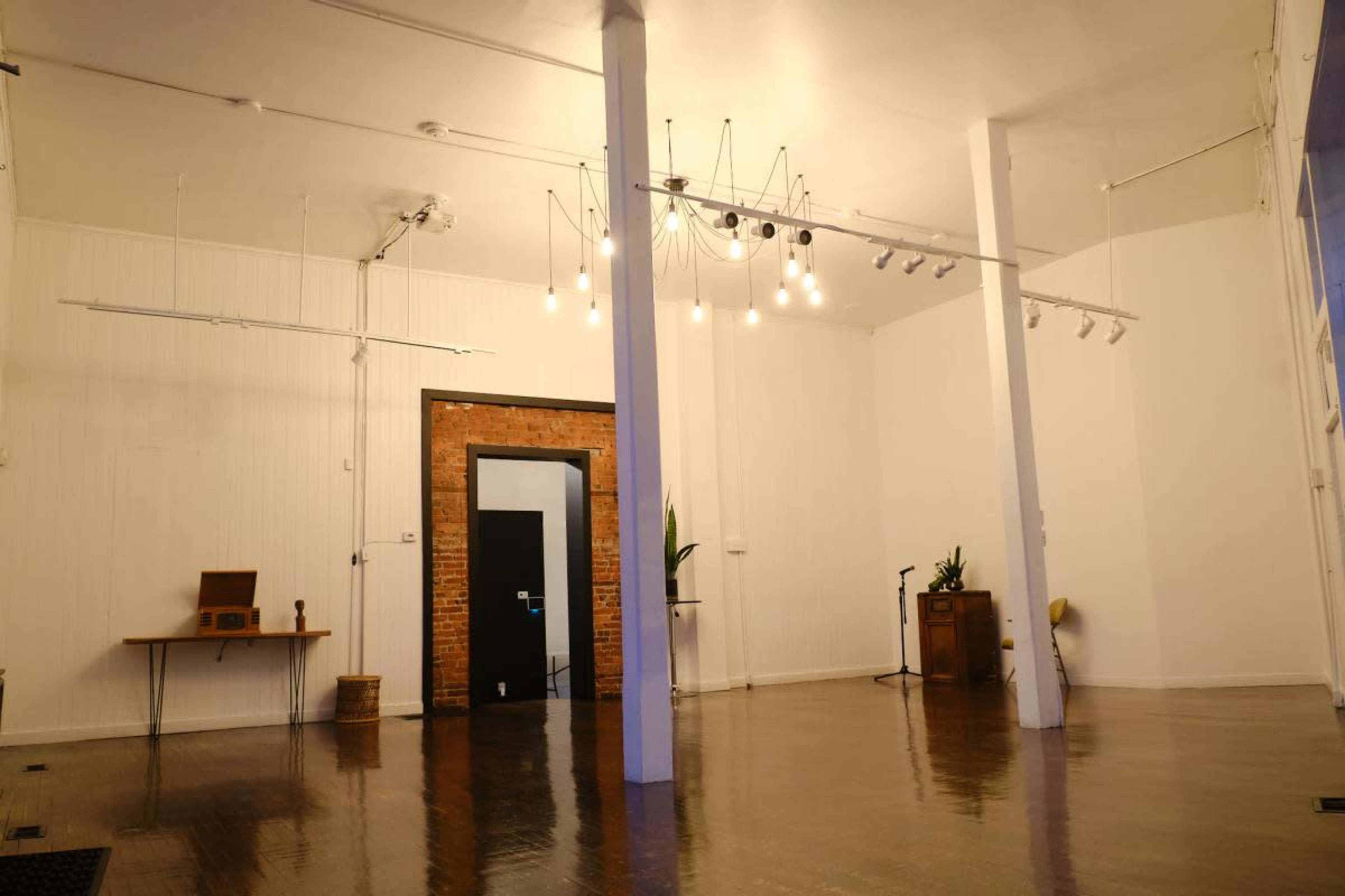 The image shows an empty, spacious room with wooden floors, white walls, and a mix of overhead lighting, including hanging bulbs and track lights.