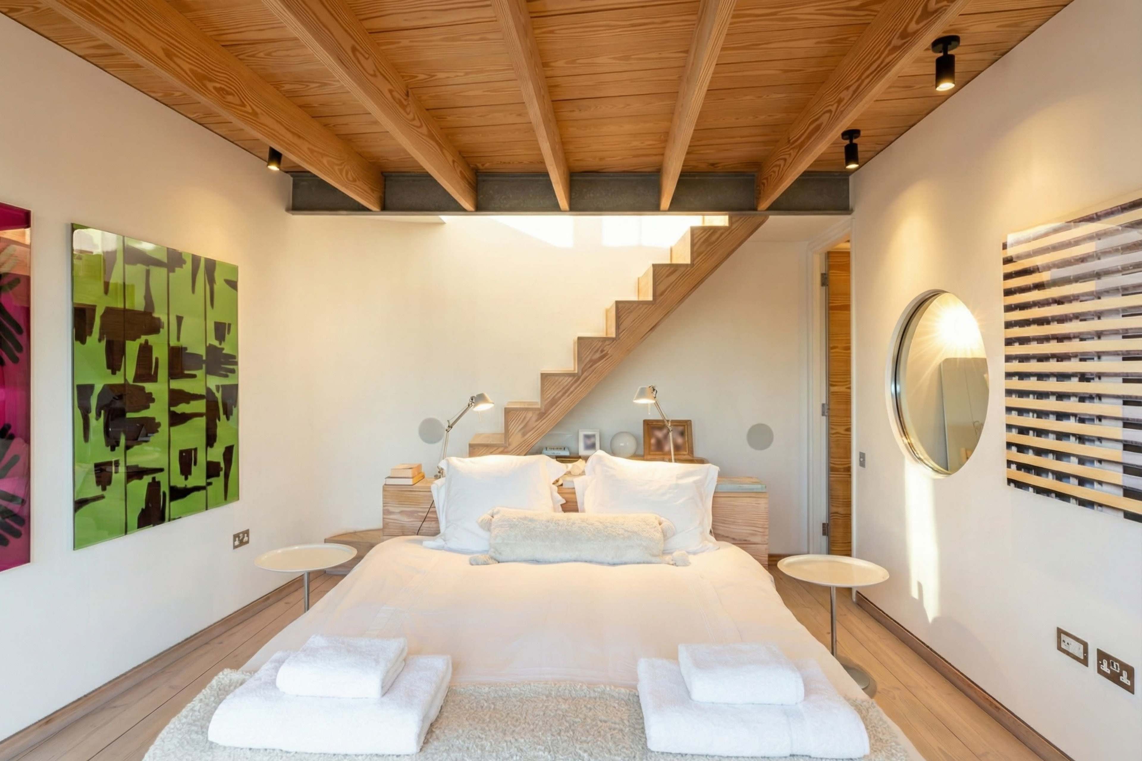 A modern bedroom features a bed with white bedding and two side tables, with a staircase and artwork on the walls.