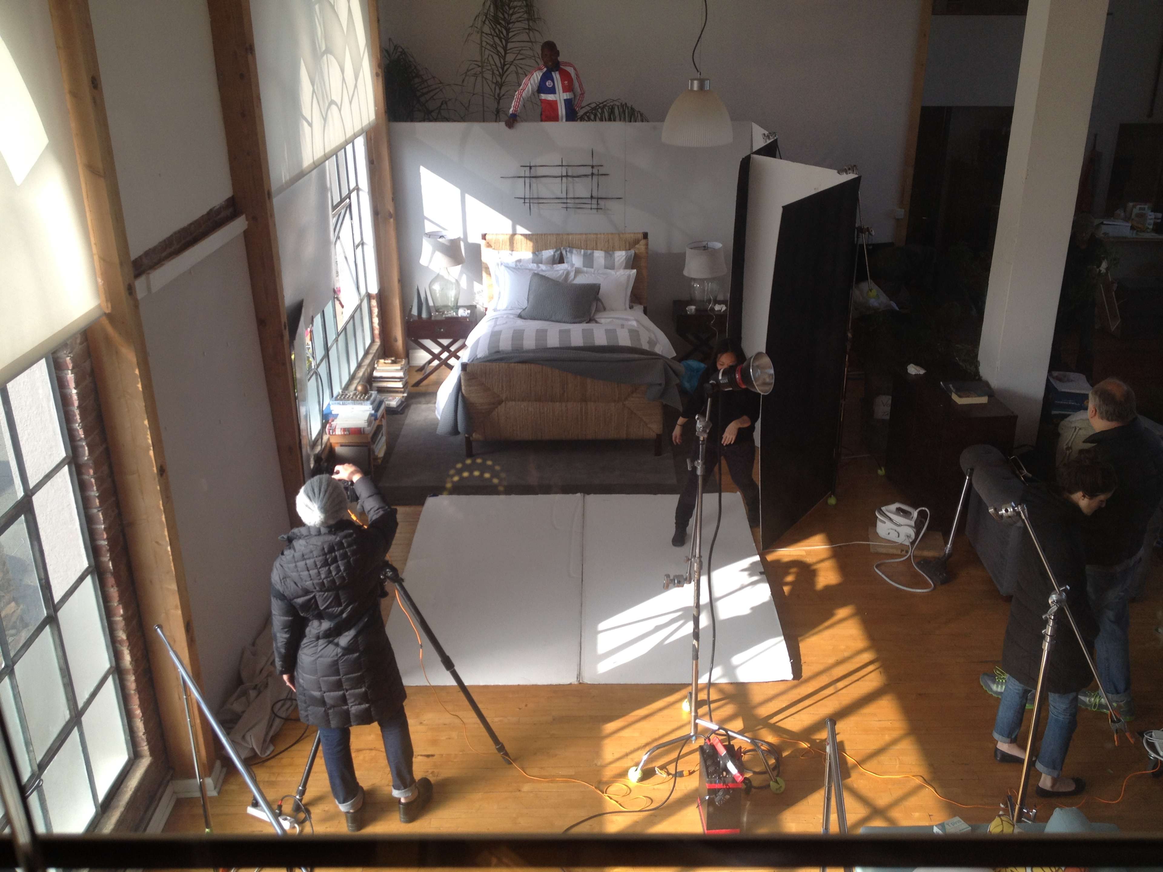 A photography shoot is taking place in a spacious room with a bed, surrounded by equipment and crew members.