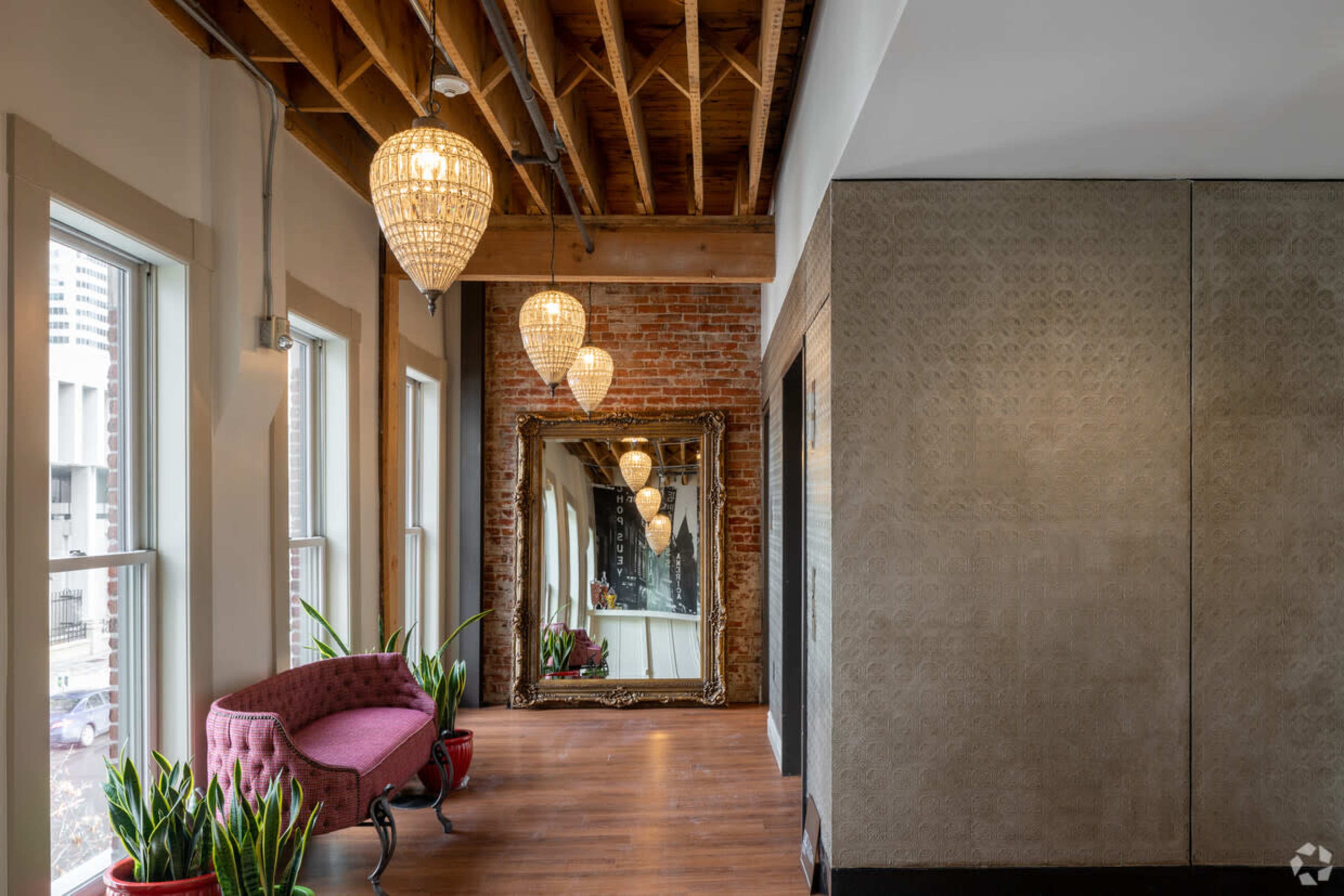 A hallway features a pink upholstered bench, decorative chandeliers, a large ornate mirror, and potted plants near the windows.