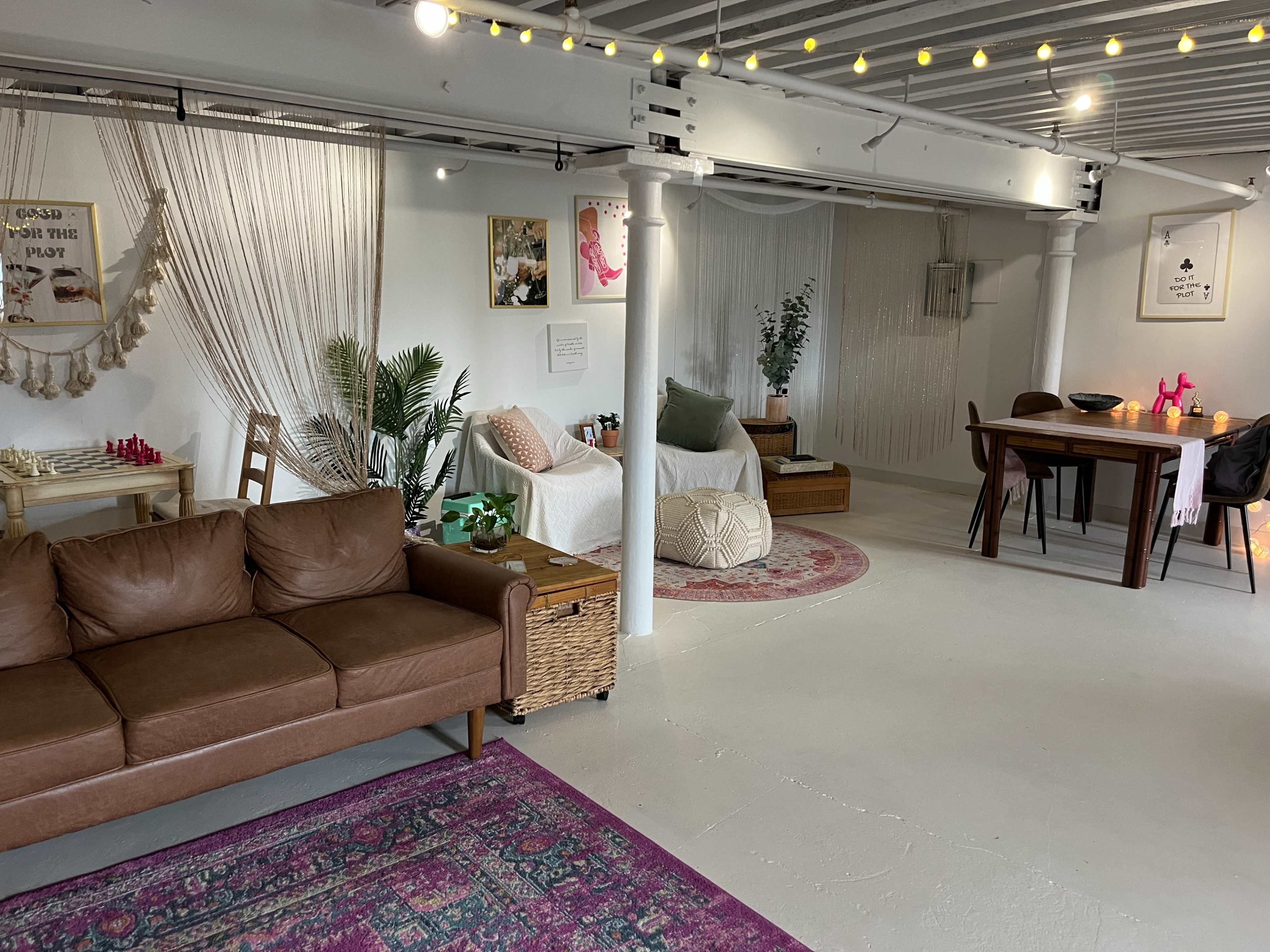 The image shows a cozy, multi-functional room with a brown leather sofa, a dining table, various seating areas, and decorative elements like plants and textiles.