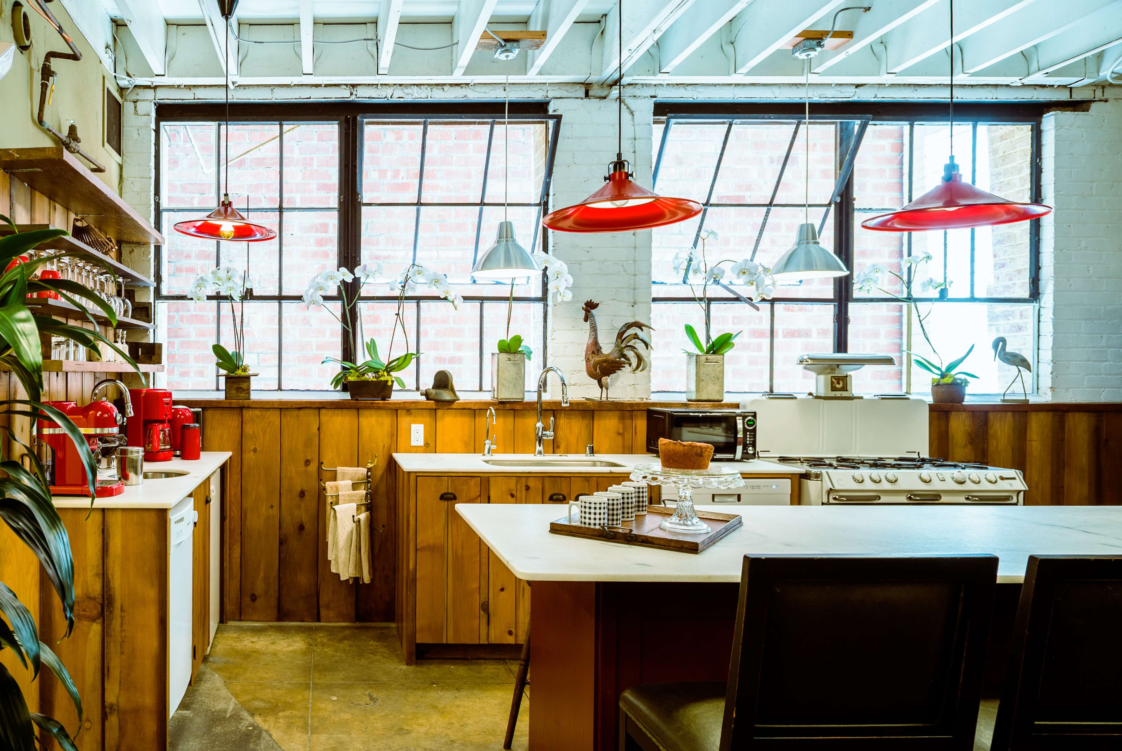 The kitchen features wooden cabinetry, a central island with seating, red pendant lights, and large windows bringing in natural light.