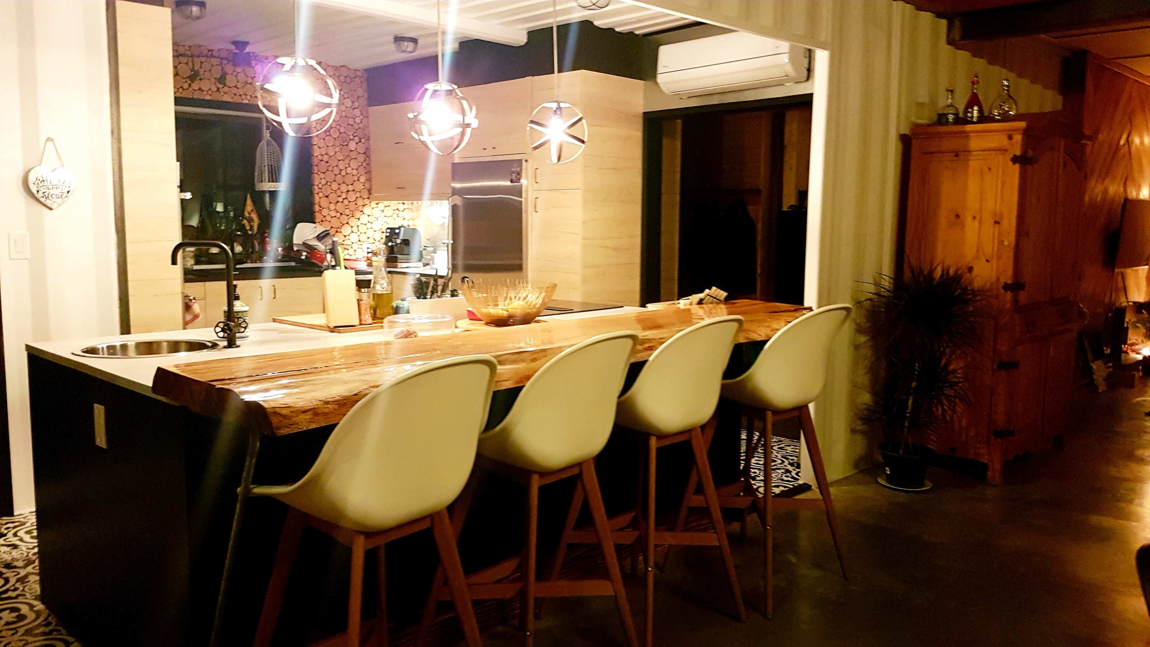 A modern kitchen with a wooden bar counter and five white chairs, featuring pendant lights above and a stylish interior design.