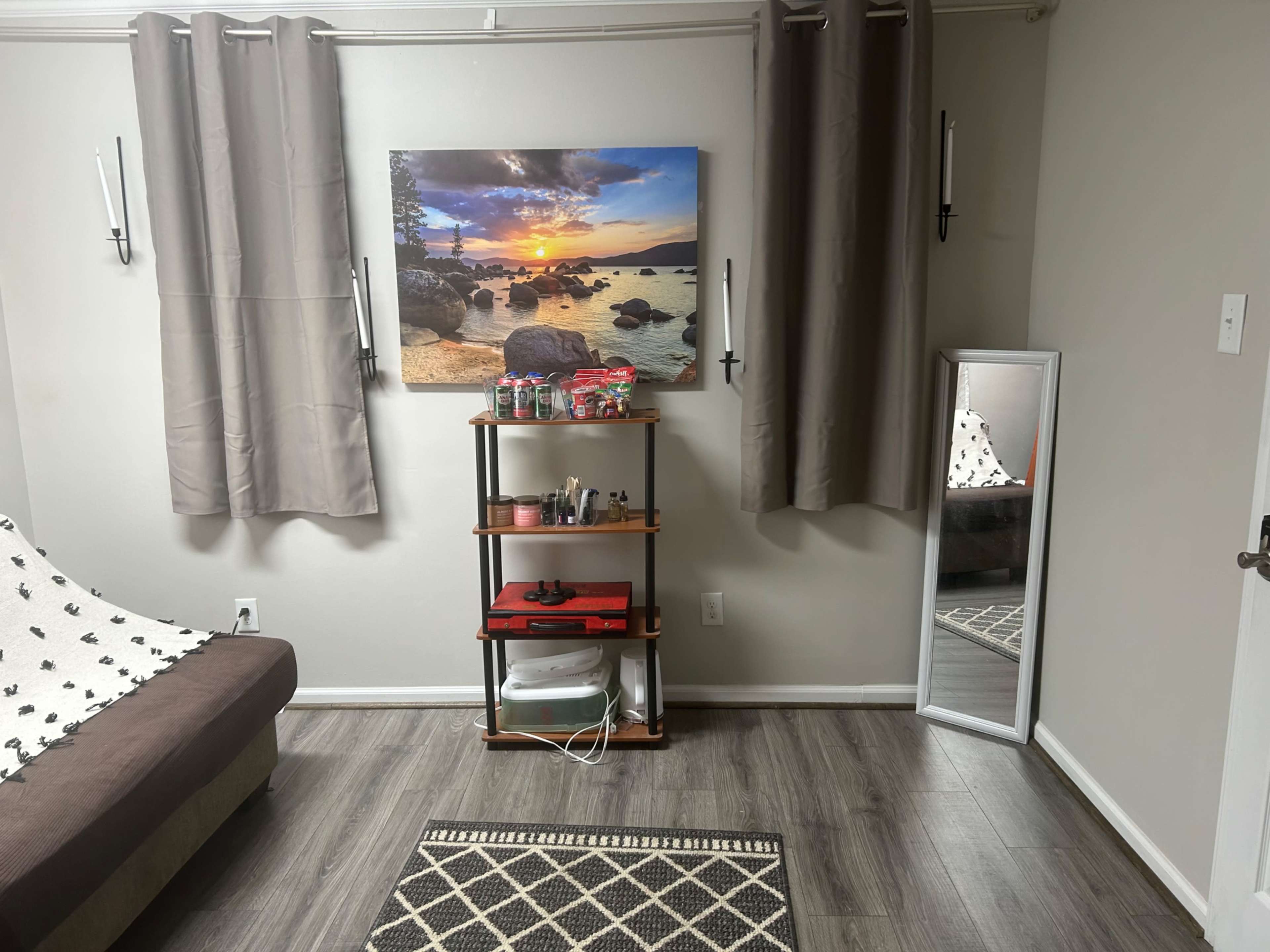 The image shows a living room with a brown couch, a shelf displaying various items, a large wall-mounted photograph of a sunset over water, and a full-length mirror next to the wall.