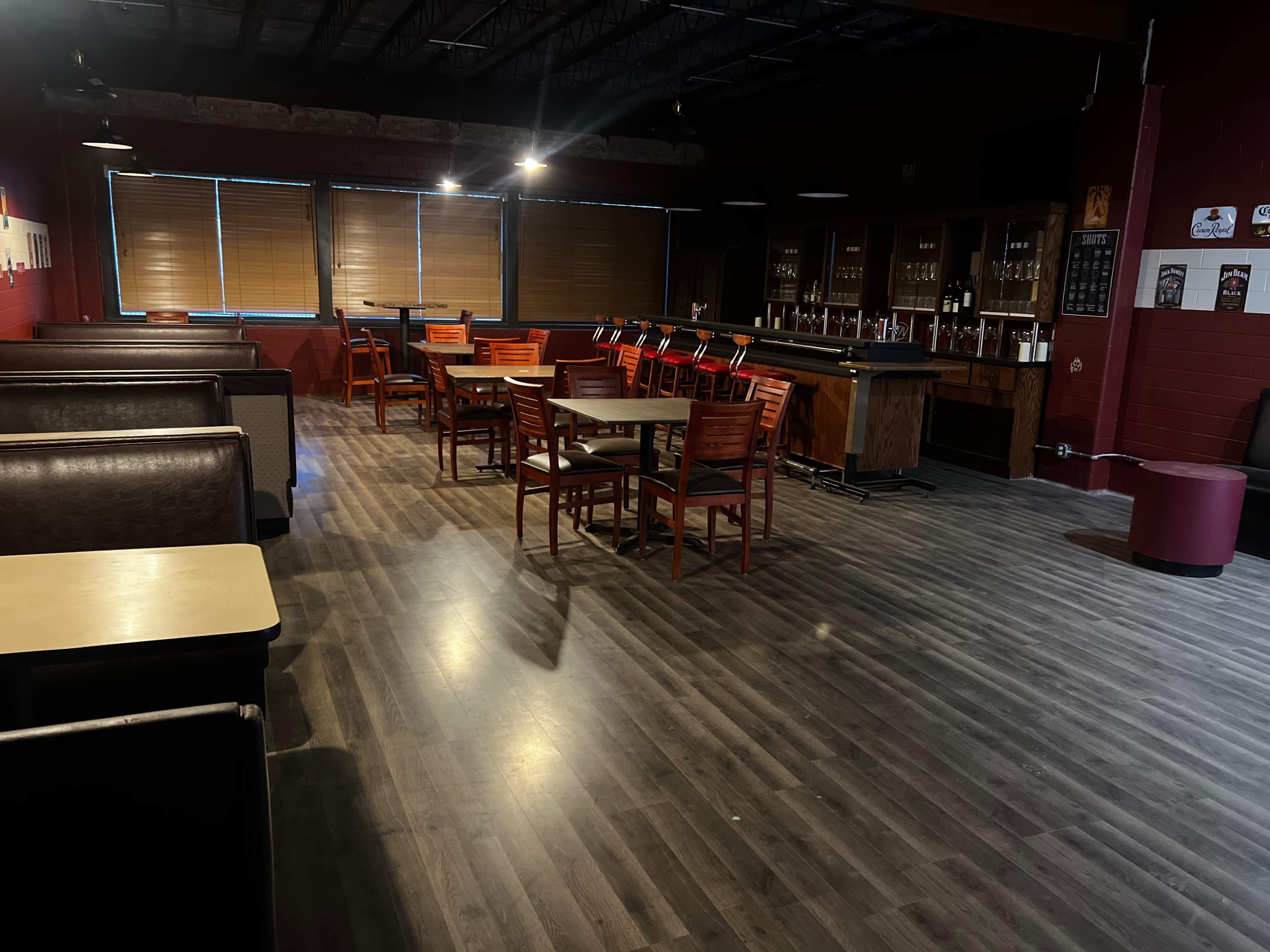 A spacious bar area with wooden tables and chairs arranged in rows, illuminated by overhead lights, and a bar counter lined with taps in the background.