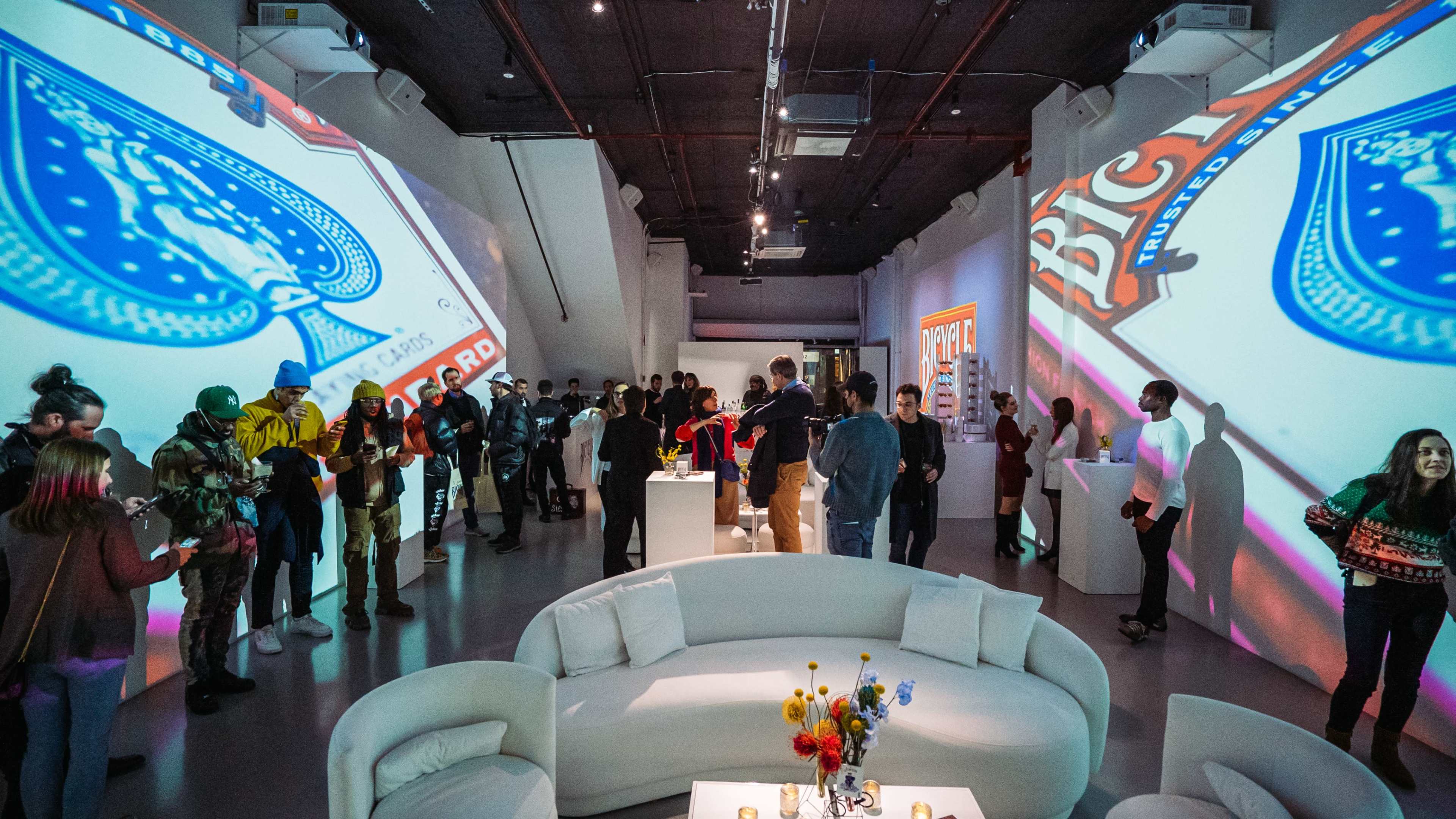 A modern gallery space filled with people interacting and colorful projections on the walls.