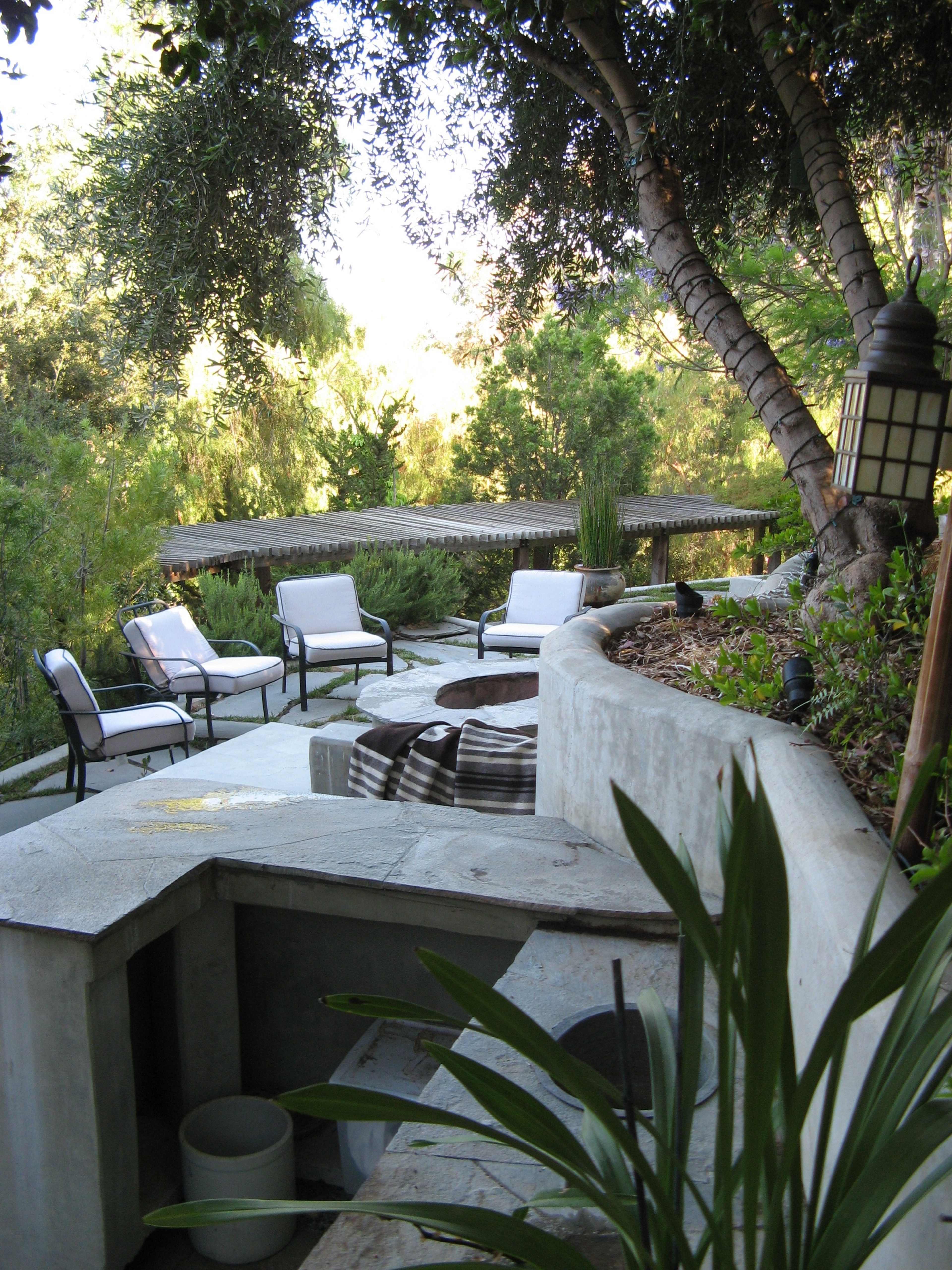 A landscaped outdoor seating area featuring several chairs arranged around a stone fire pit, with greenery and trees in the background.