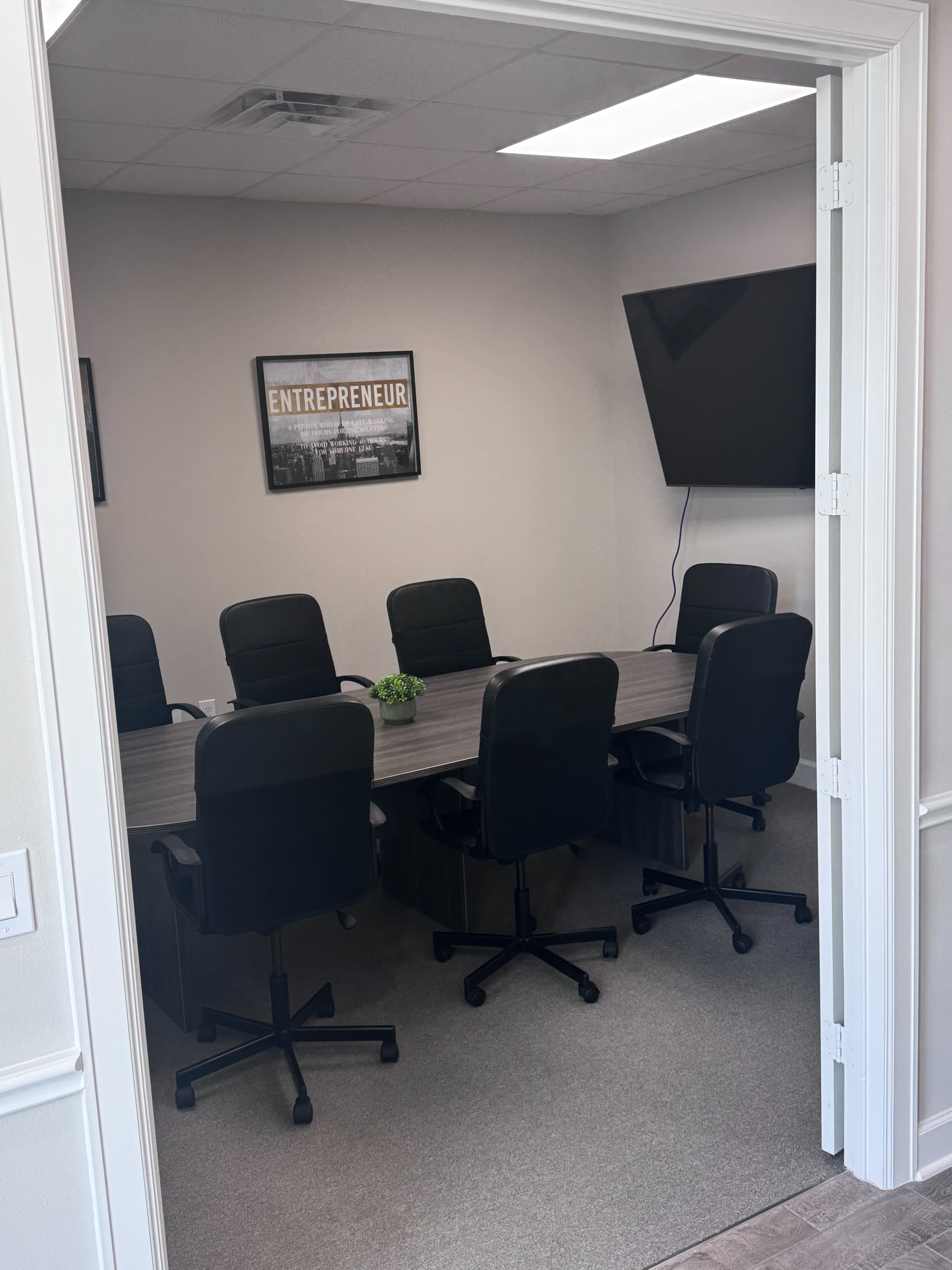 The image shows a conference room with a large table surrounded by six black swivel chairs and a wall-mounted television.