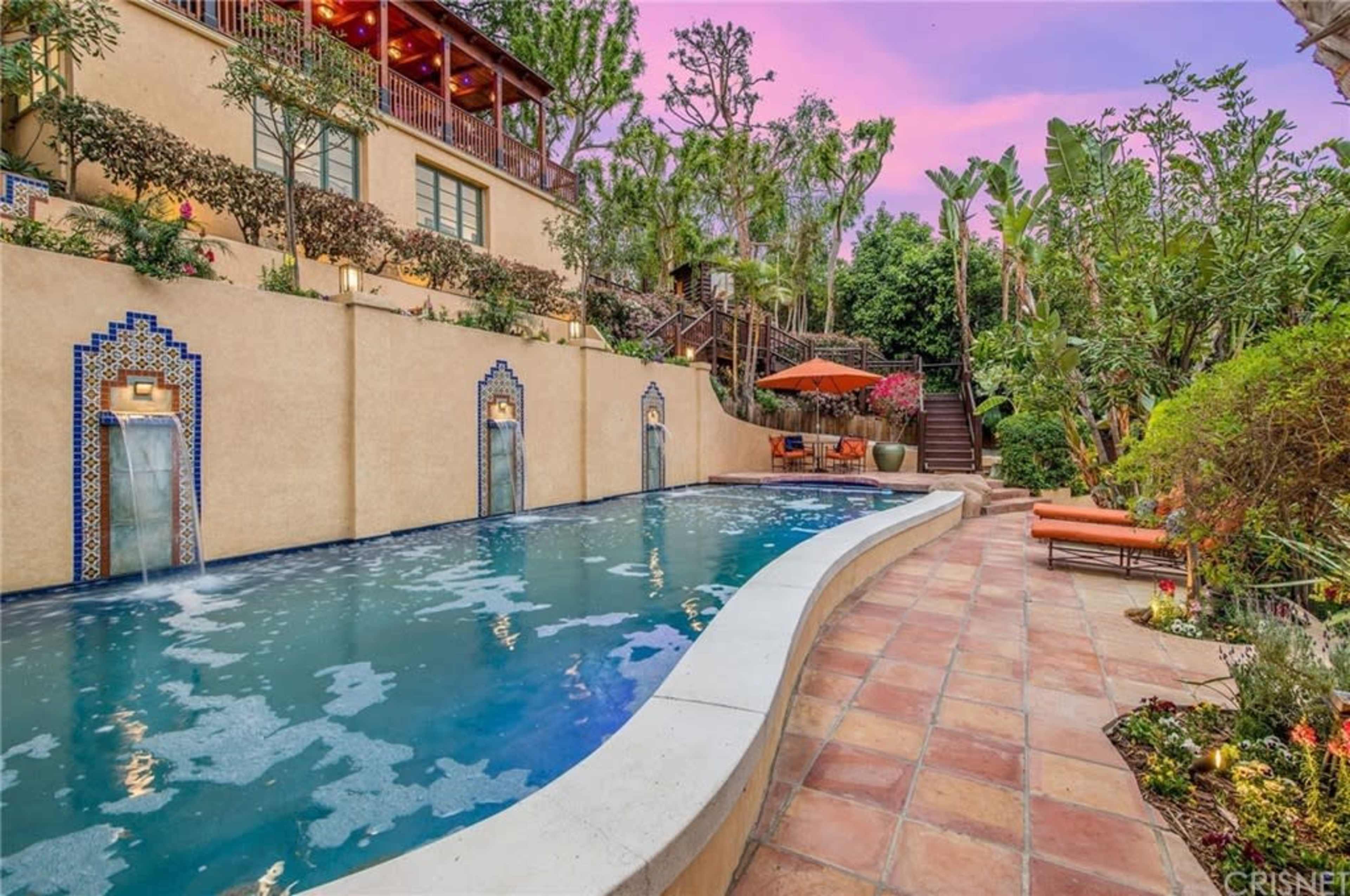 A landscaped backyard featuring a curved swimming pool bordered by a stone patio, with lounge chairs, stairs, and greenery surrounding the area.