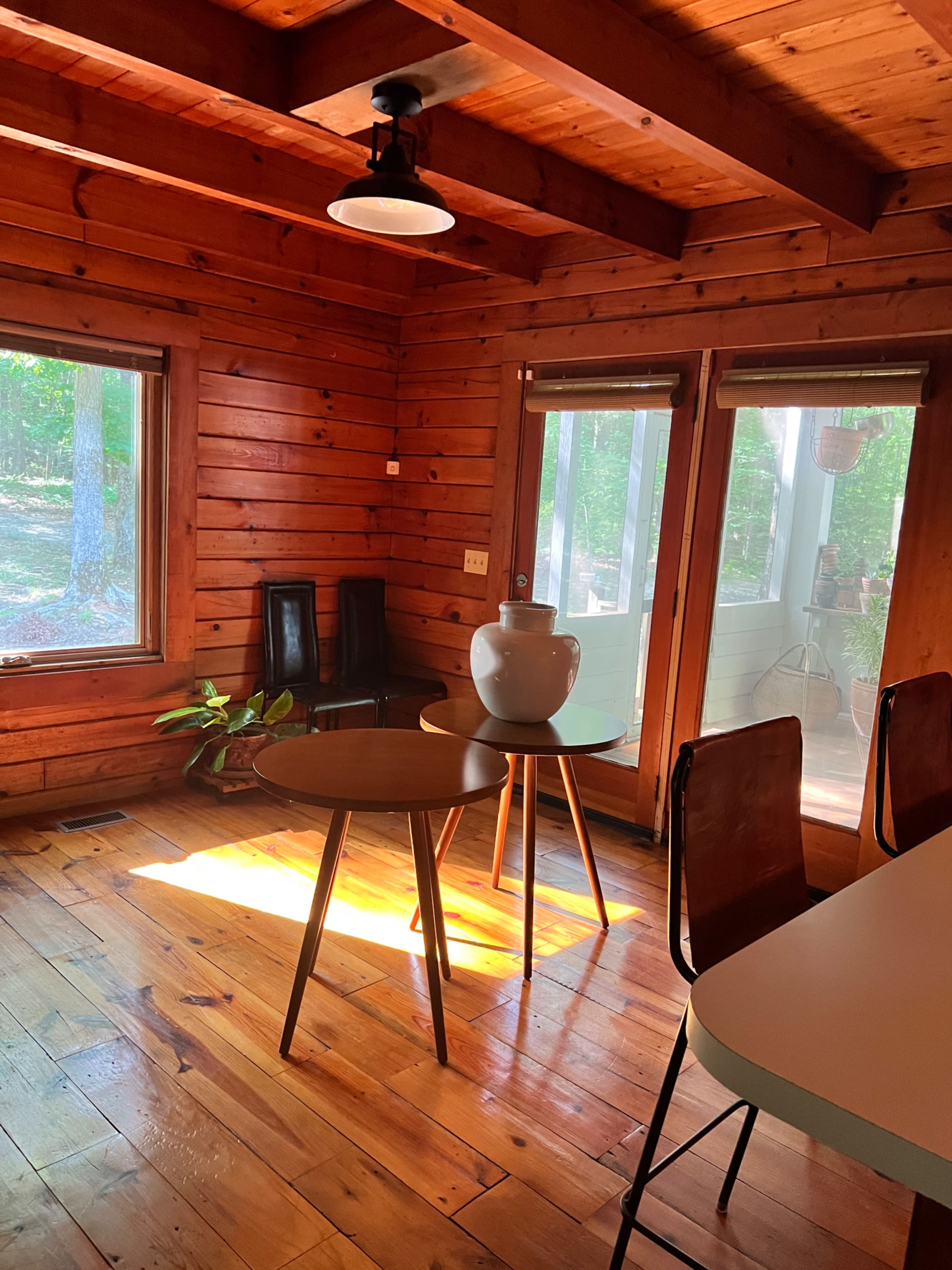 The room features wooden walls and floors, with a small round table and two black chairs near a large window allowing sunlight to enter.