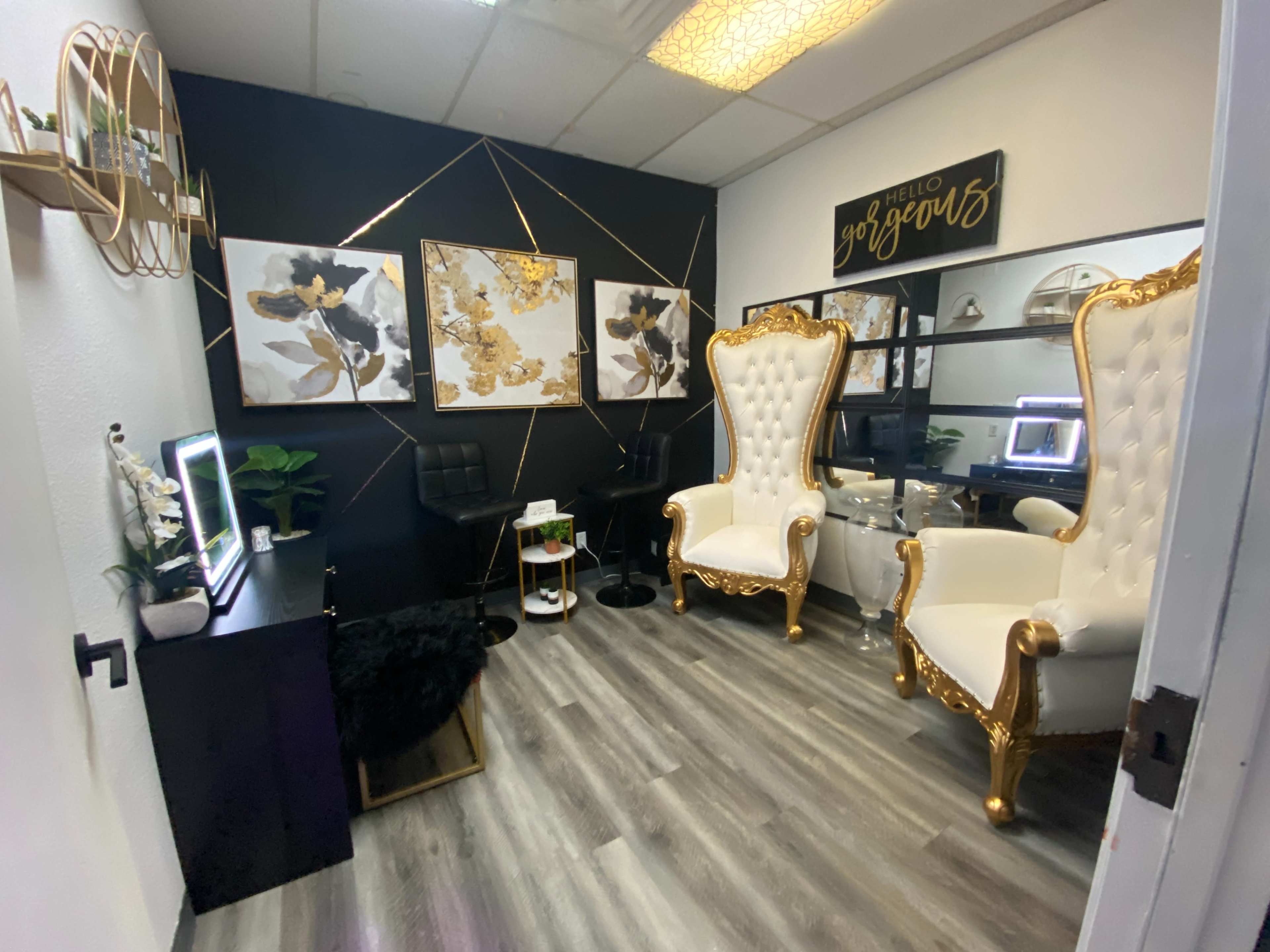 A small, stylish beauty studio with two ornate white and gold chairs, black walls adorned with art, and a neatly arranged workspace.