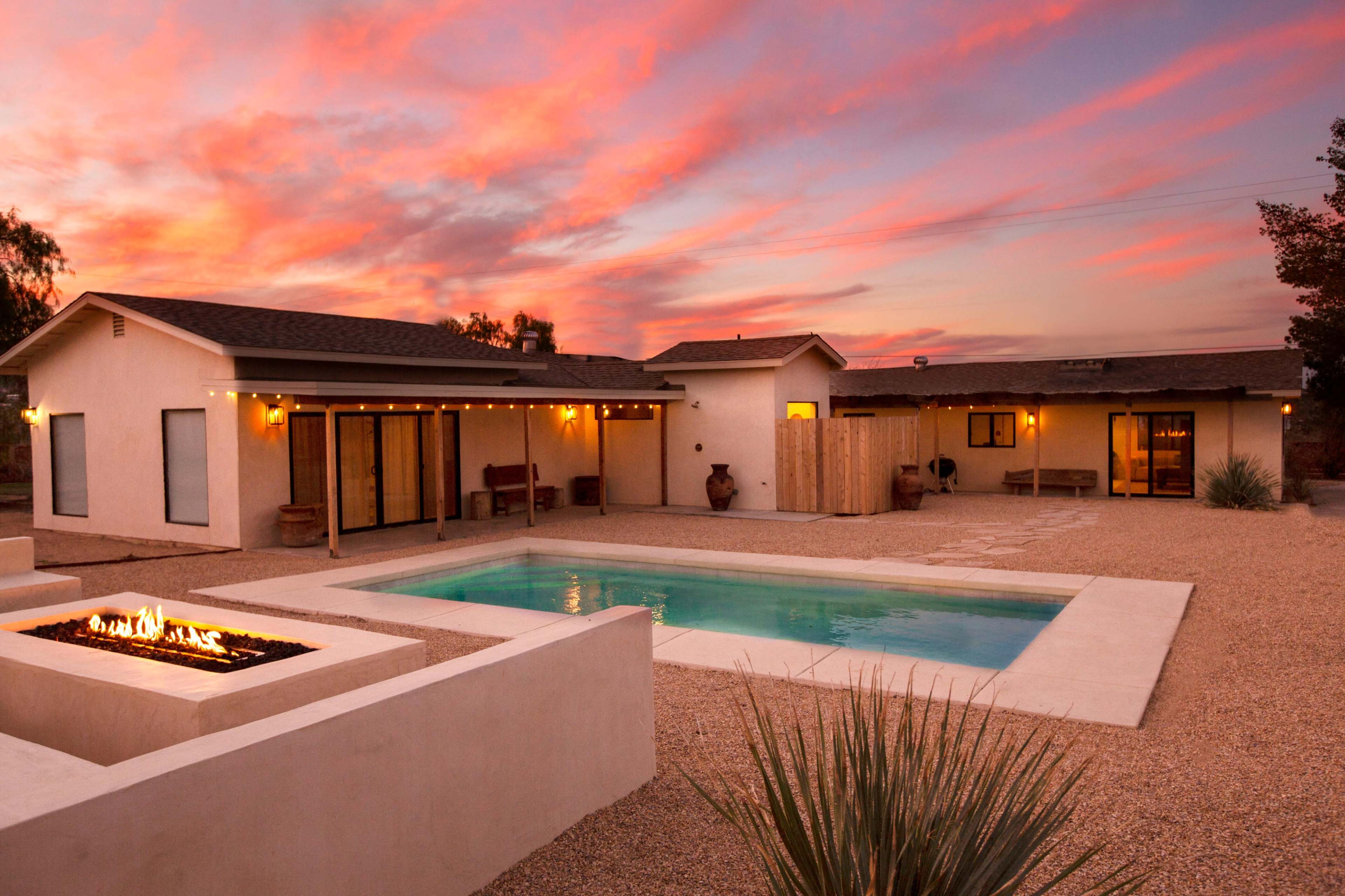 A modern home with a pool and fire feature is surrounded by a gravel courtyard under a sunset sky.