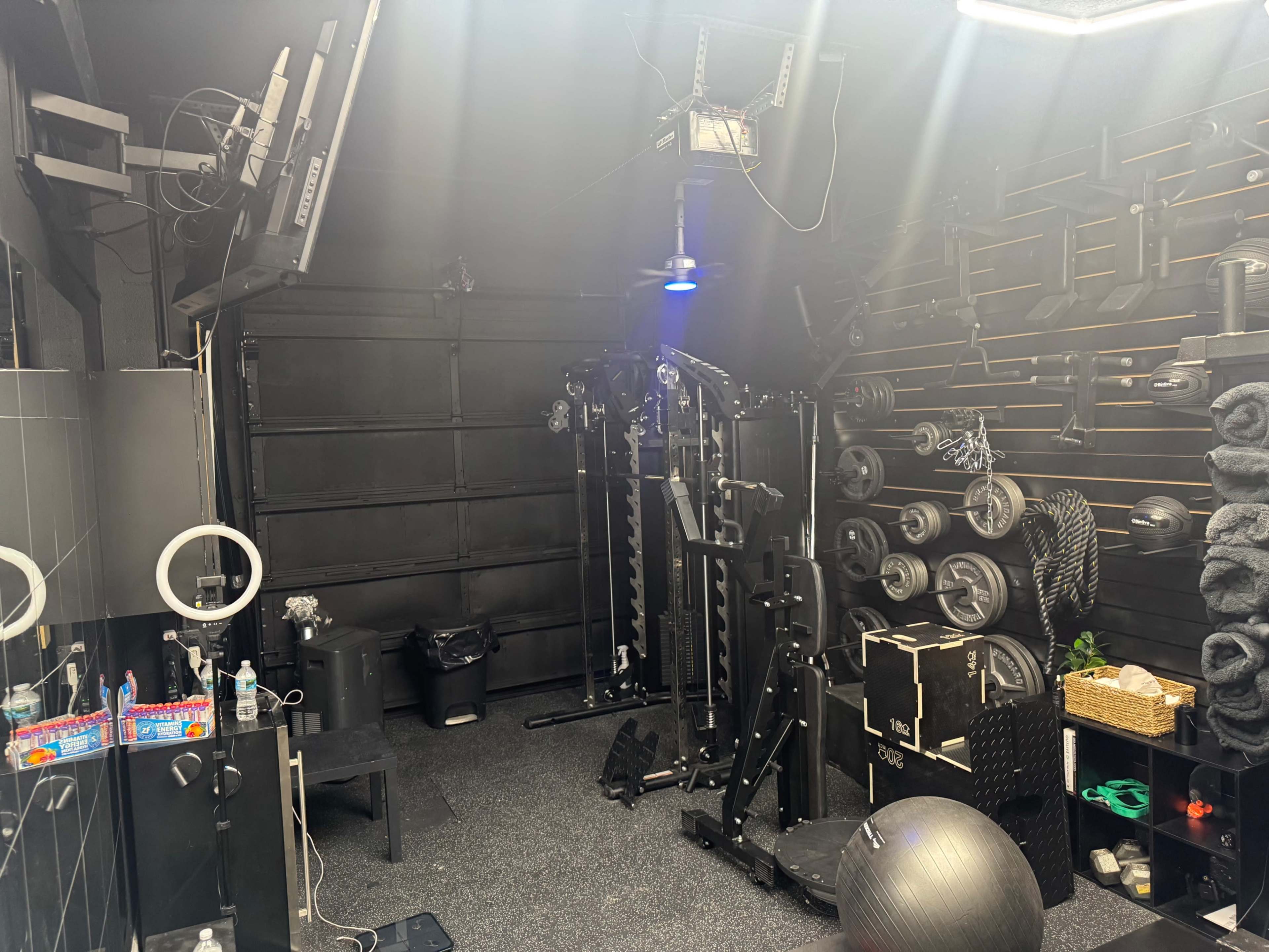 The image shows a compact home gym with various weightlifting equipment, a bench, mirrors, and floor mats arranged against dark walls.