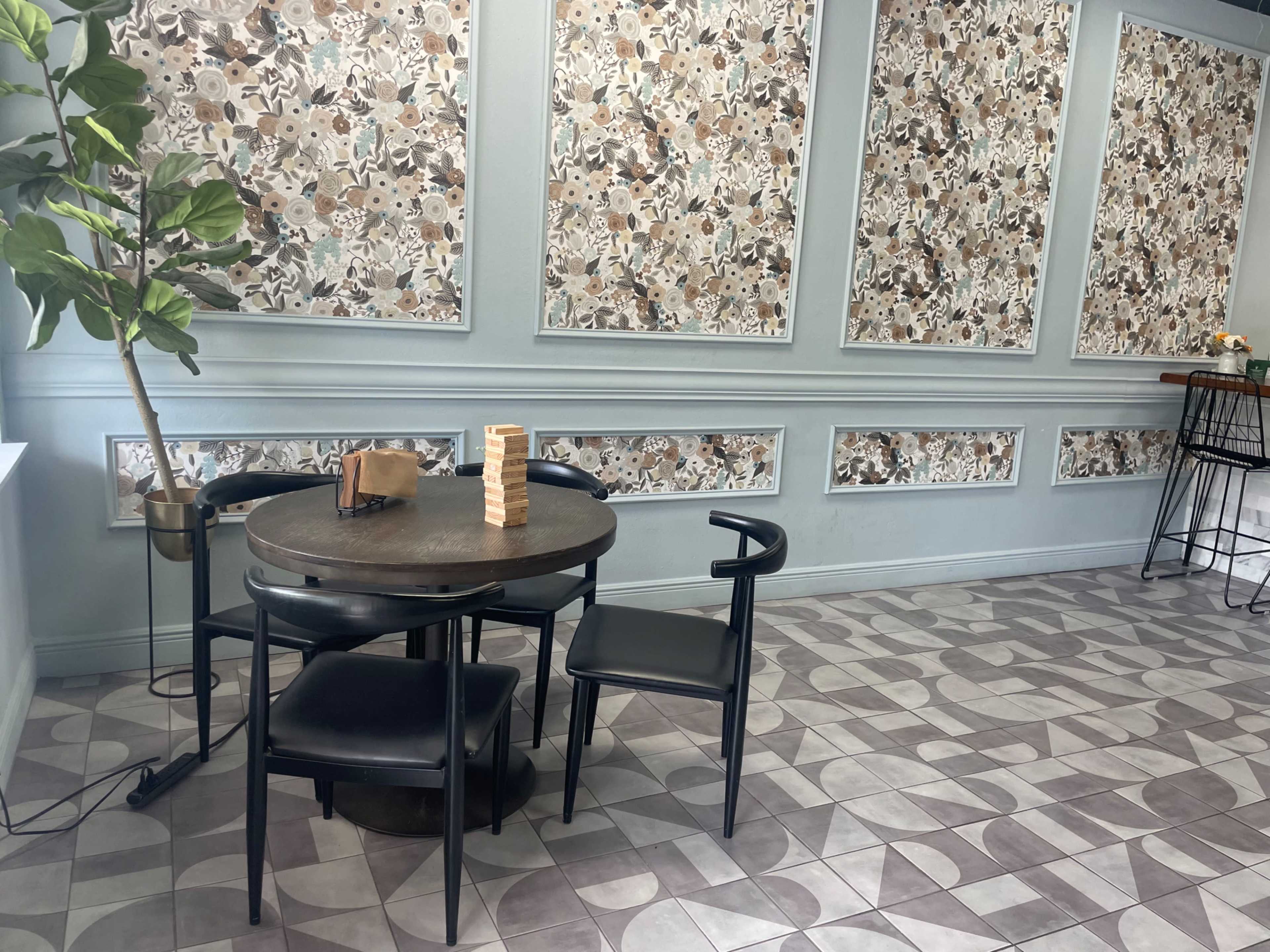 A round table with black chairs is set in a cafe featuring floral wallpaper and patterned flooring.